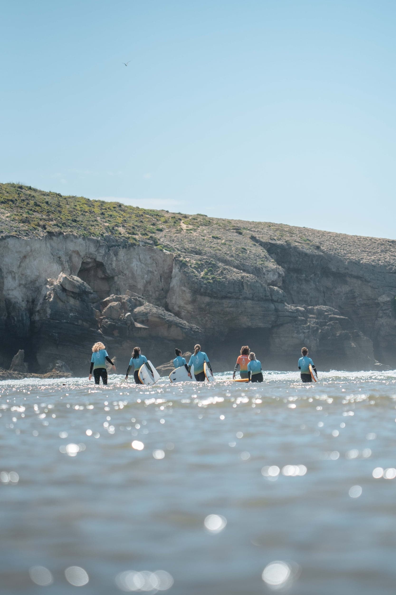 Group of surfers in blue rash vests wading out toward a cliffed cove