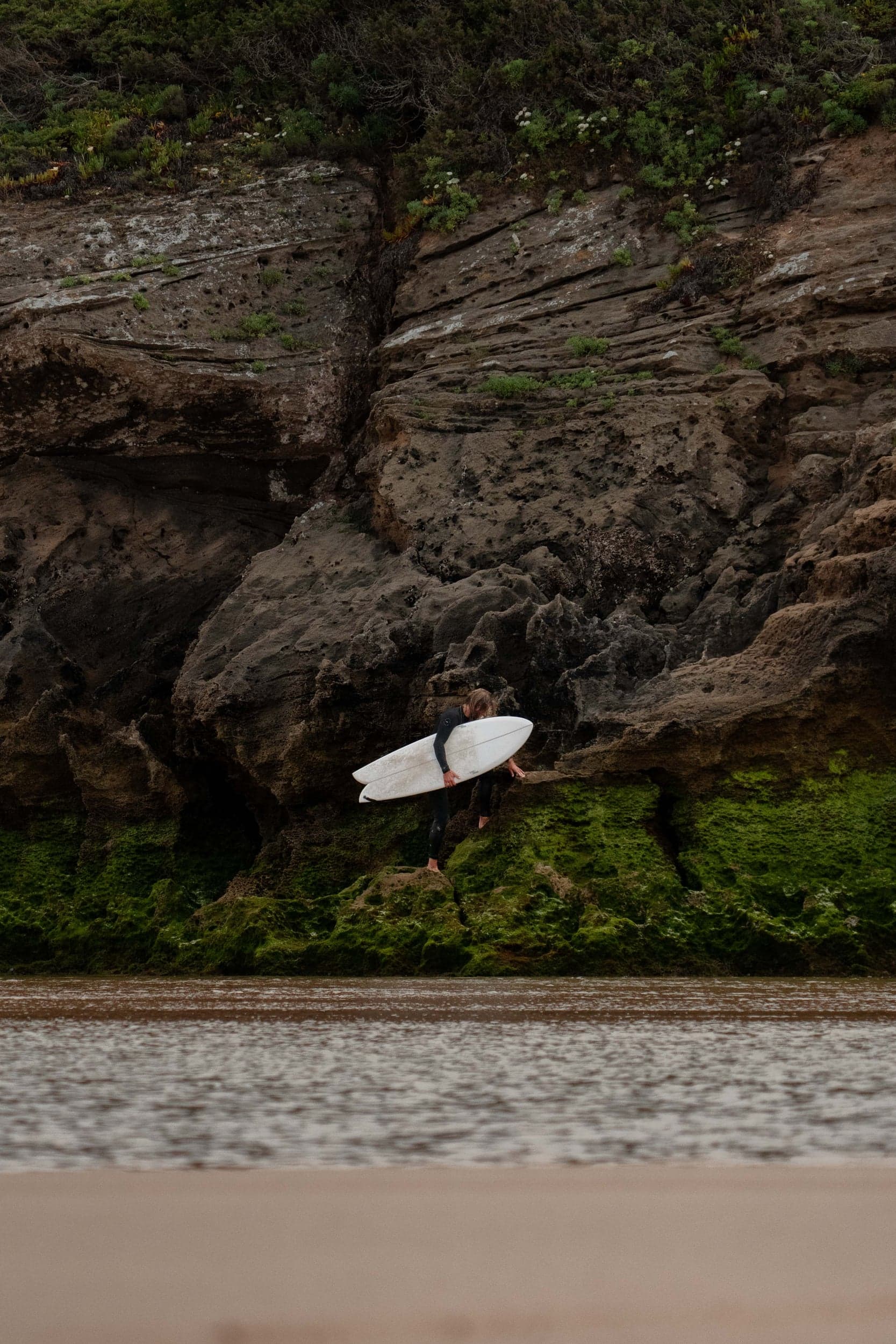 Surfer picking his way over mossy cliff rocks with a white board