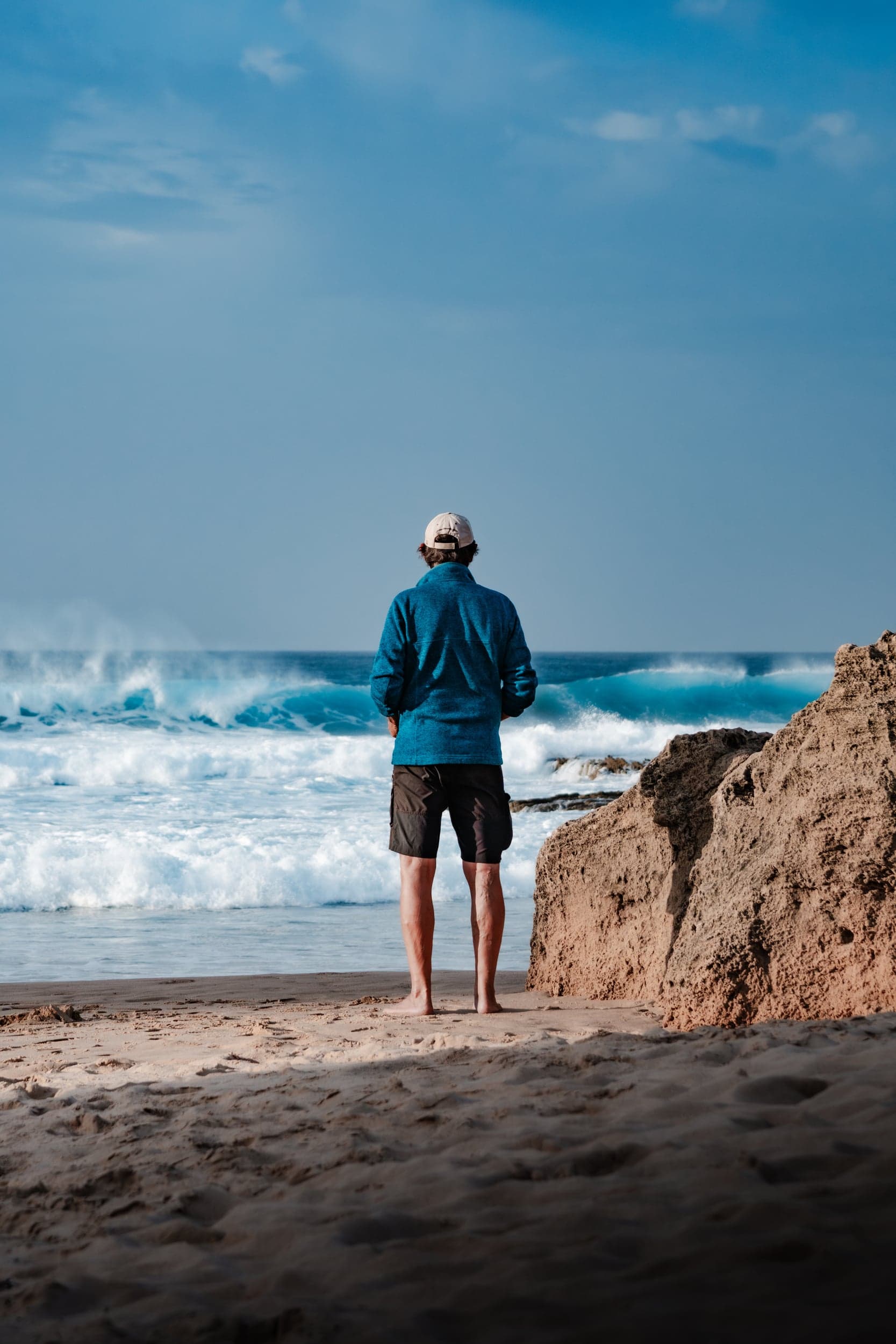 Man in a blue fleece watching the surf break from the sandy beach, back to camera
