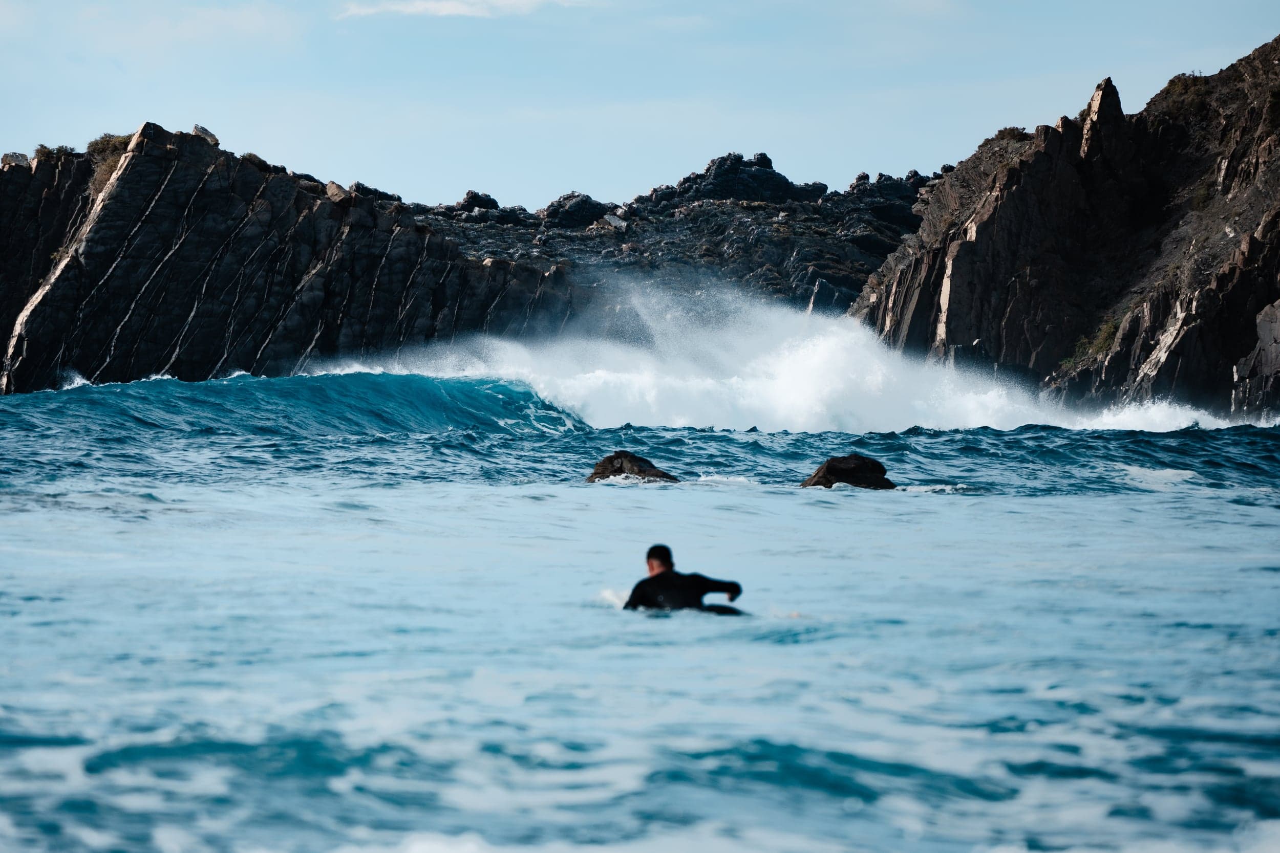Surfer paddling out toward waves breaking against jagged Algarve cliffs