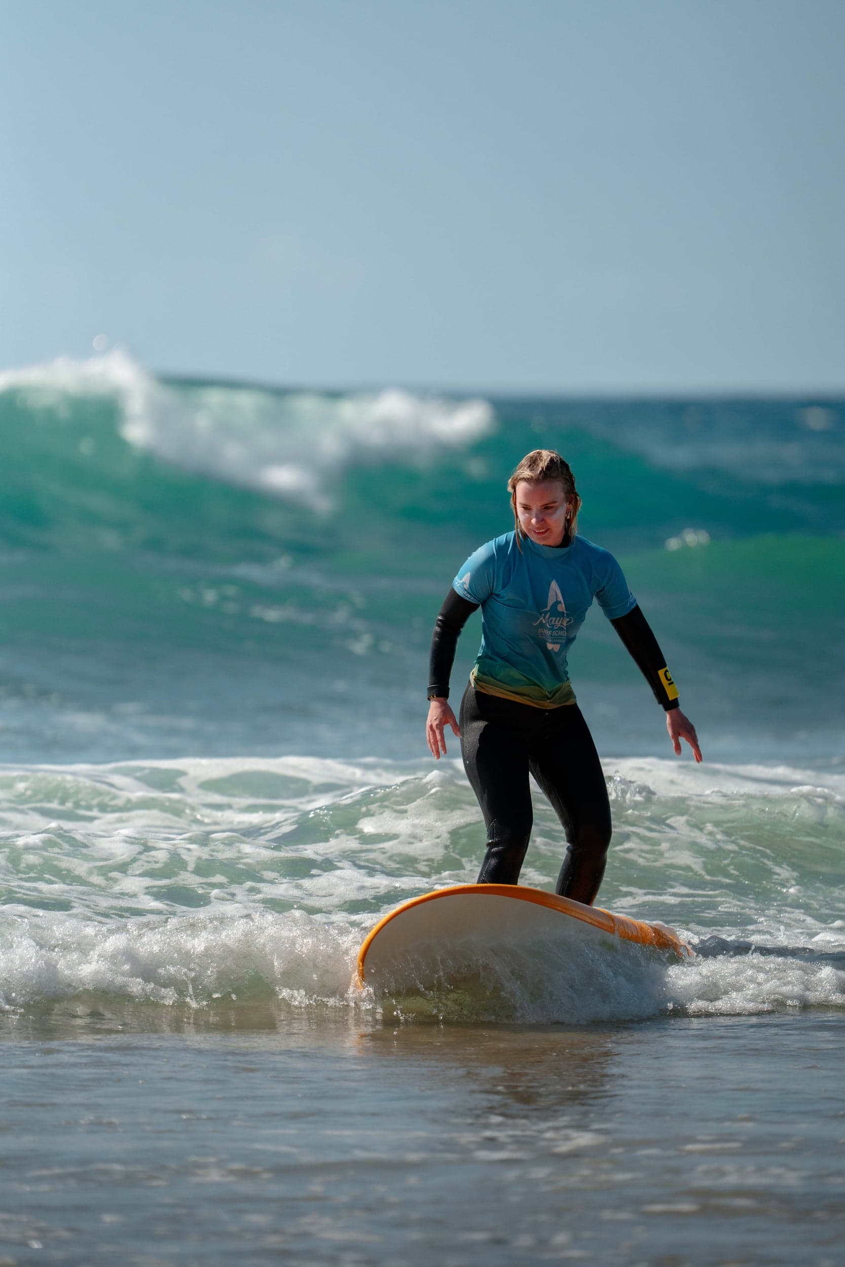 Beginner in a blue rash vest riding a foamboard through whitewater, wave breaking behind