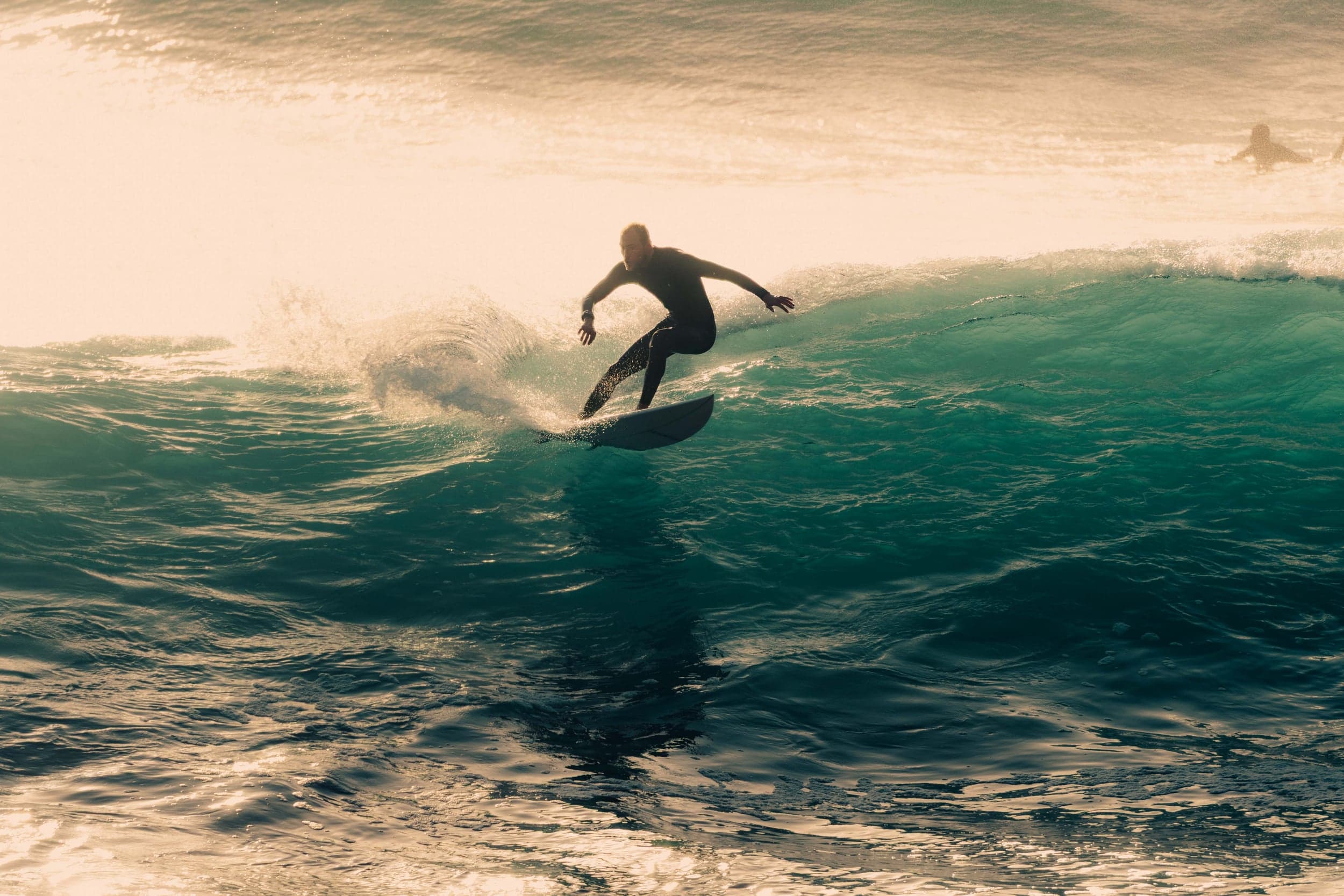 Surfer silhouetted mid-line on a glowing teal wave at sunset, spray fanning behind