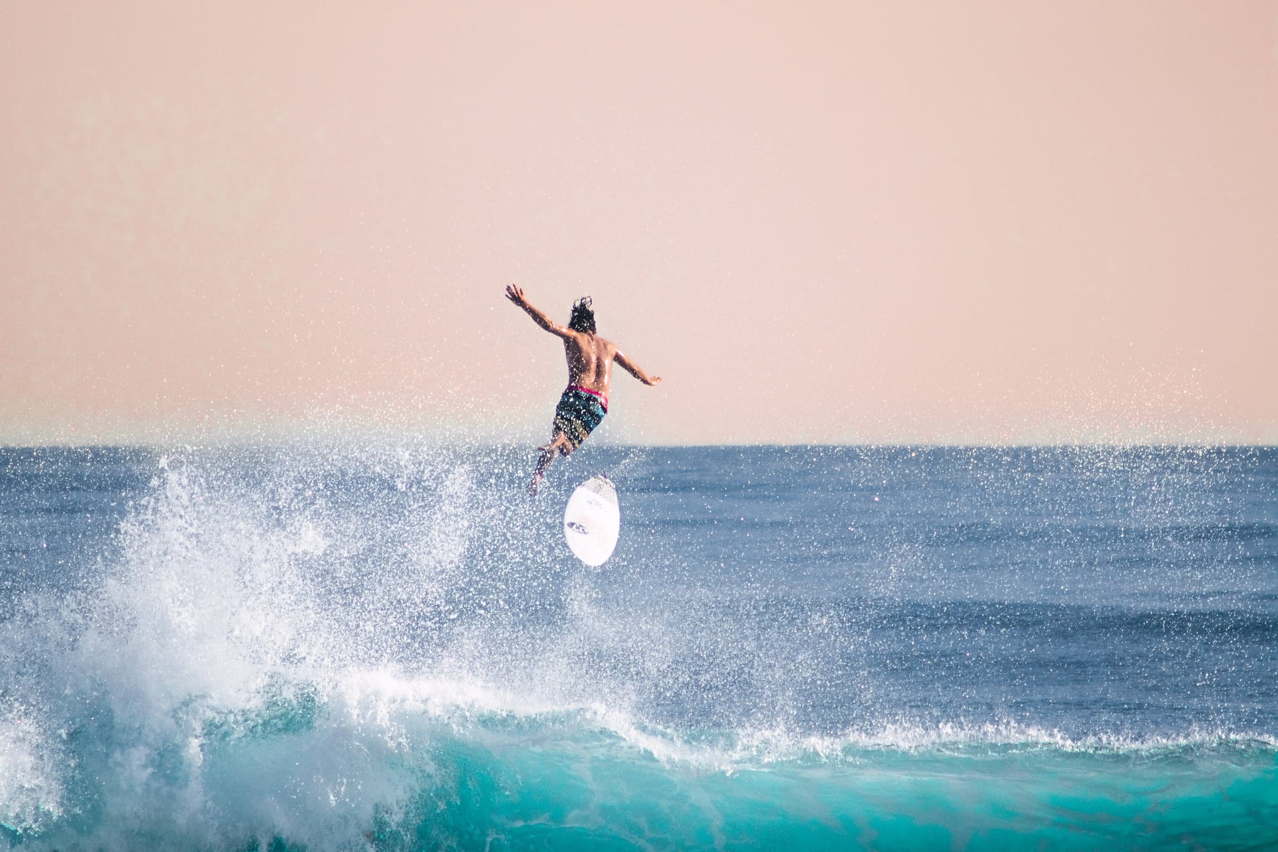 Surfer thrown from the board mid-air above turquoise water against a soft pink sky
