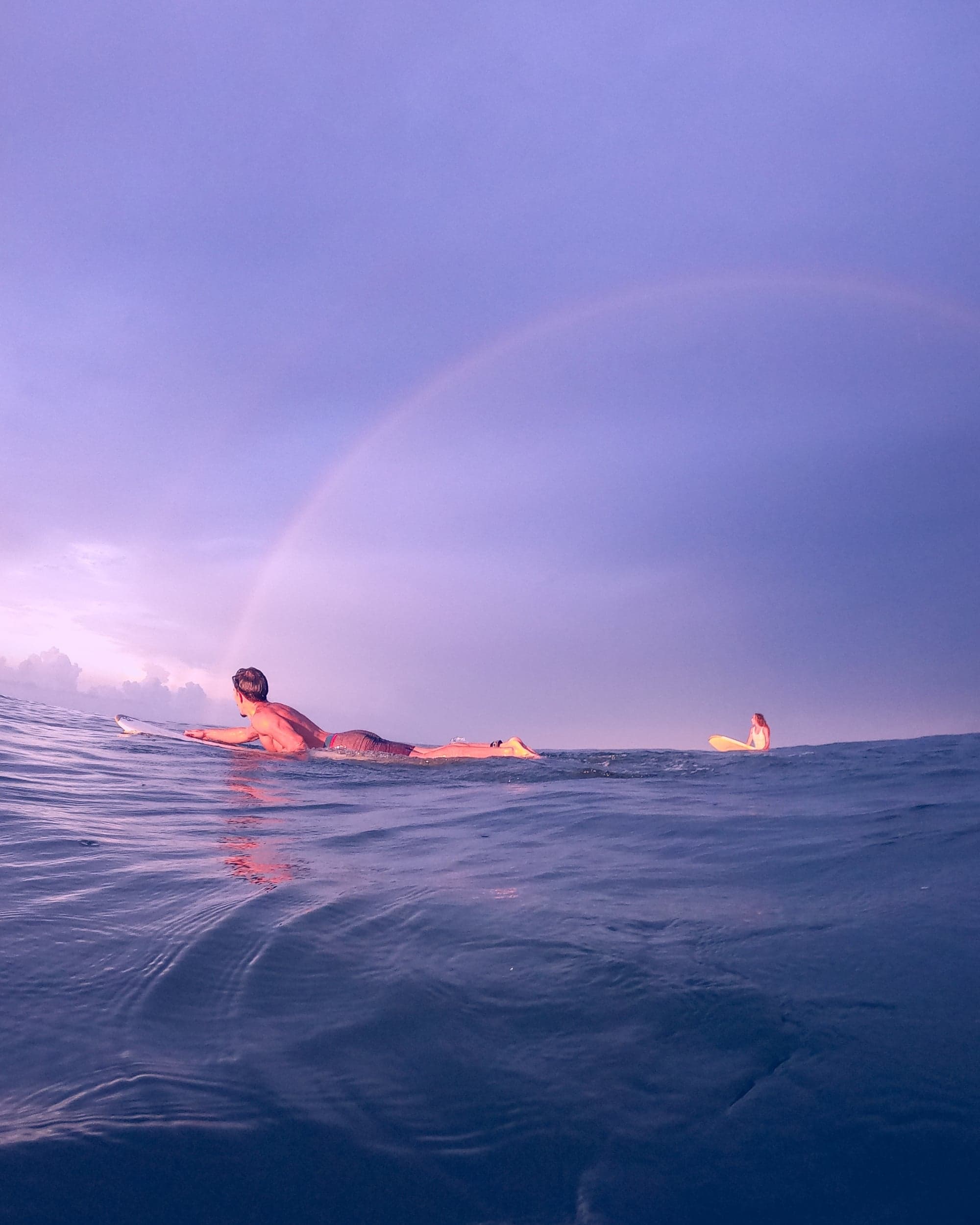 Two surfers paddling out under a faint rainbow in violet dawn light