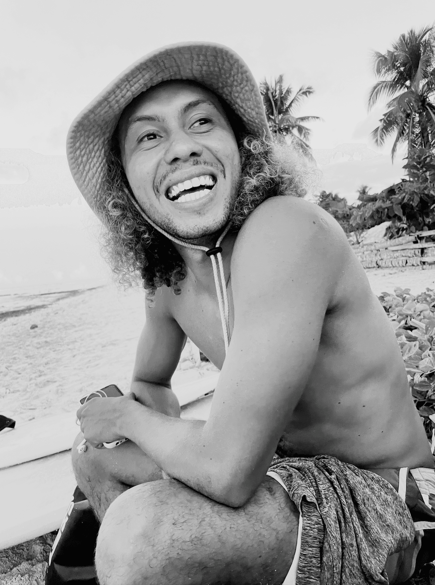 The photographer laughing in a bucket hat beside a surfboard on a palm-lined beach