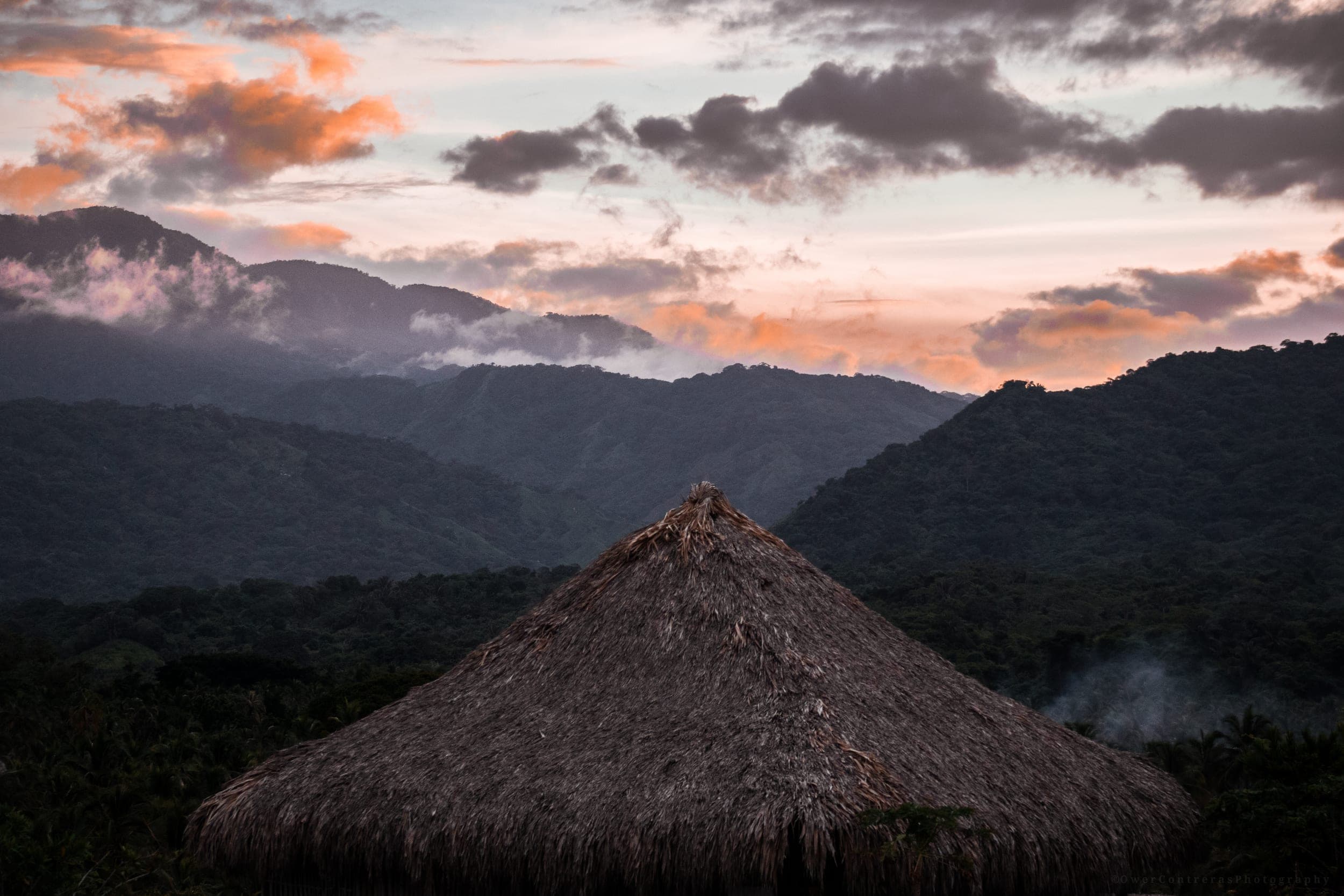 Thatched hut roof against misty jungle mountains under a soft sunset sky