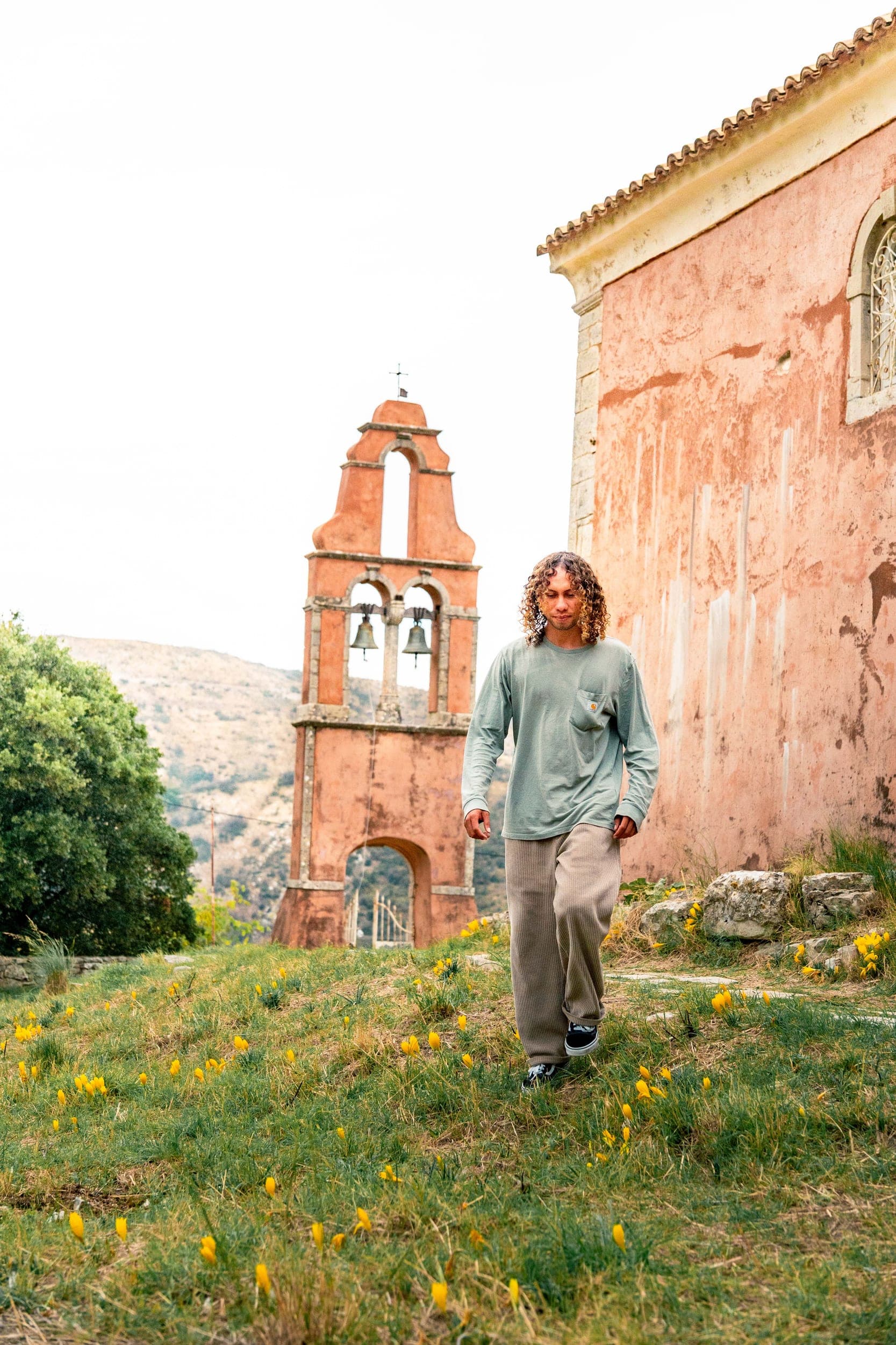Curly-haired man walking through wildflowers past an old bell tower and pink church