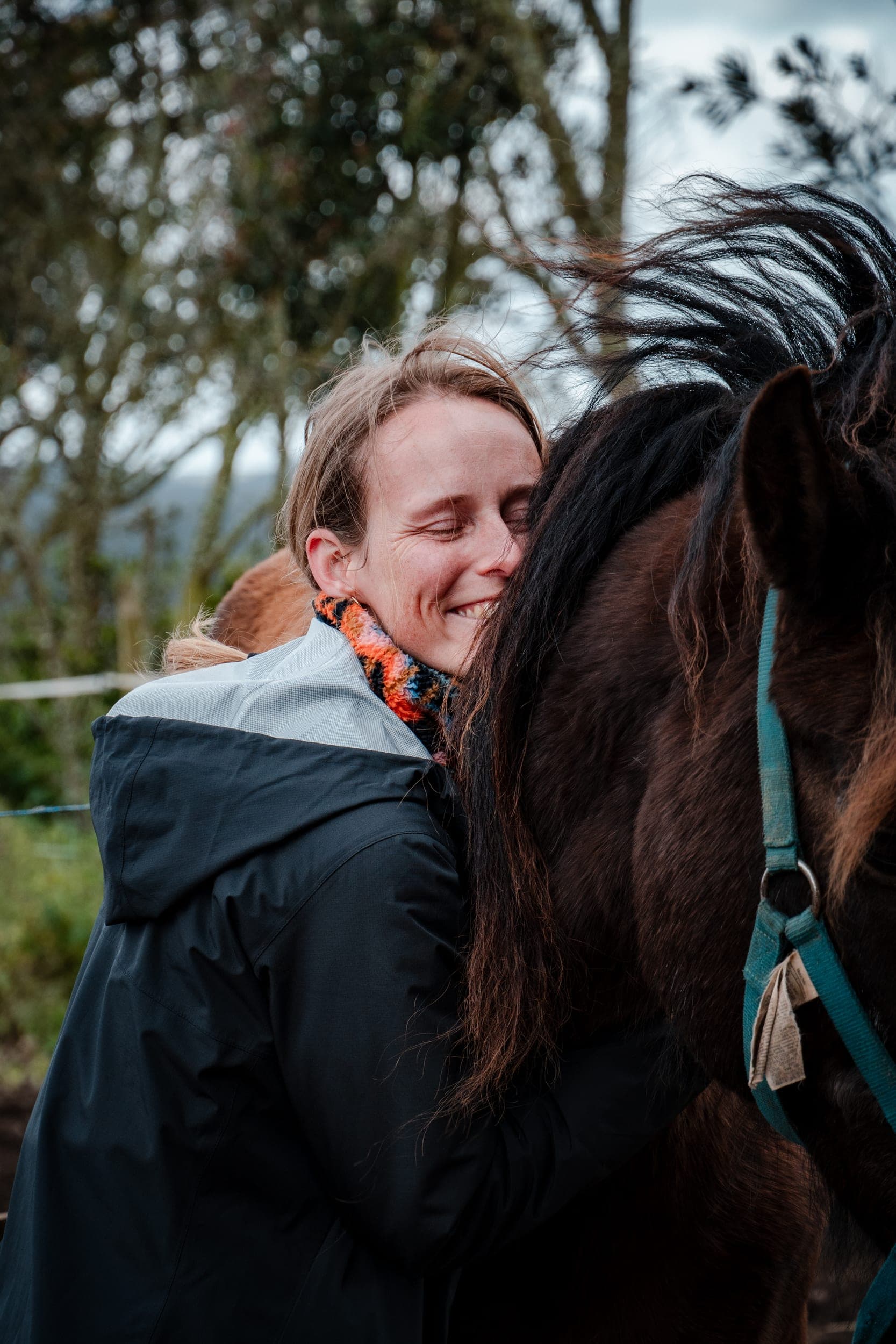 Smiling woman resting her head against a brown horse's neck at a countryside paddock