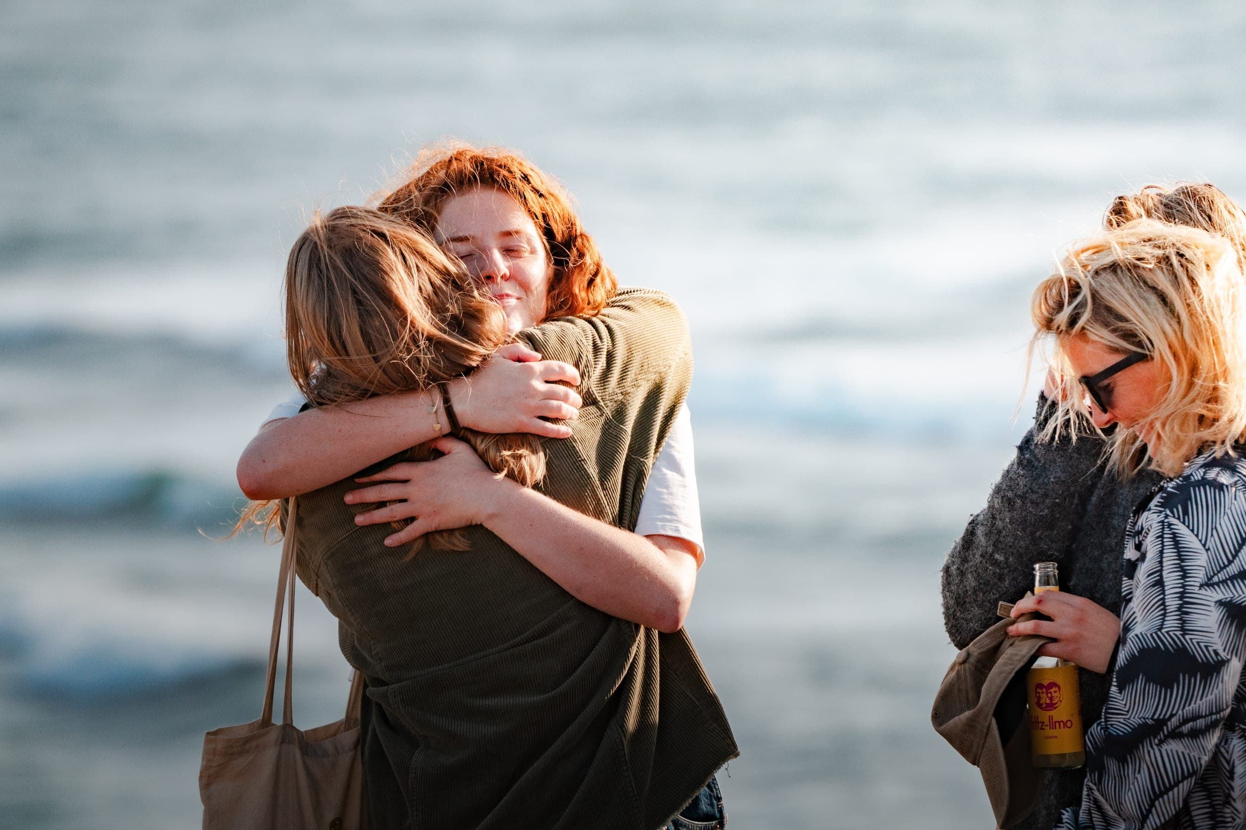 Two women hugging at sunset on the beach as a friend stands nearby holding a drink