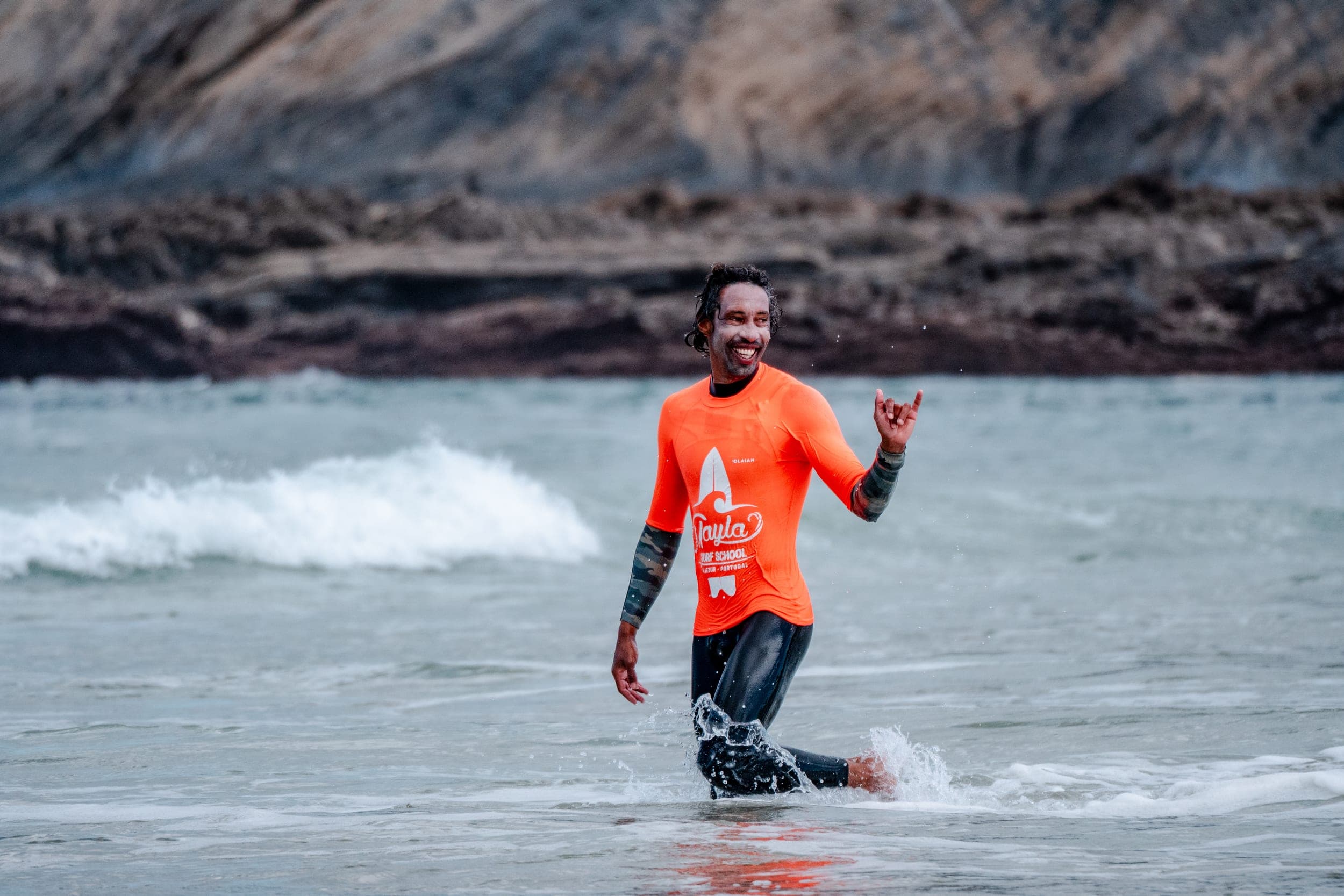 Smiling man in orange rash vest wading through shallow surf, throwing a shaka sign