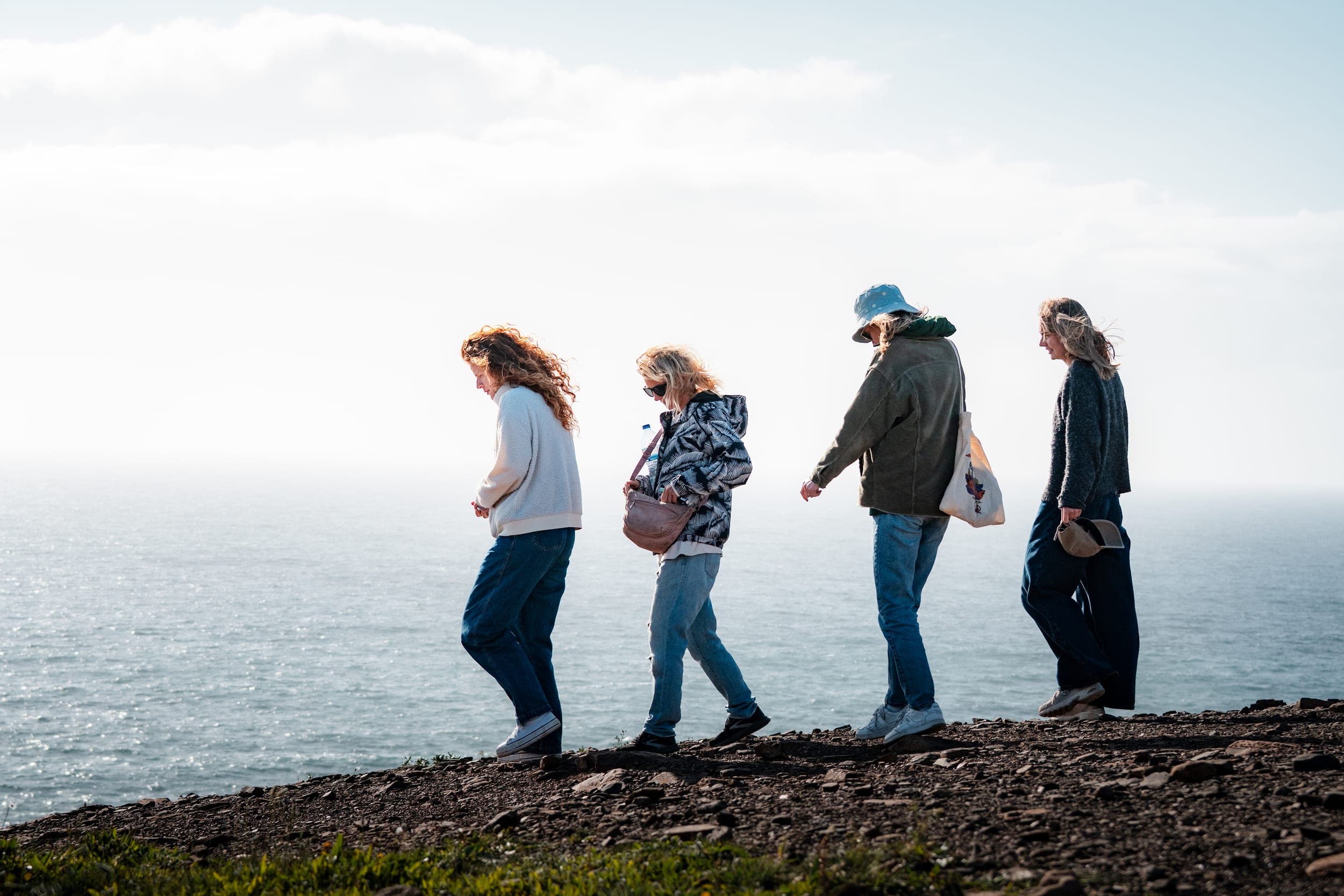 Four women walking single file along a clifftop path above a sunlit ocean