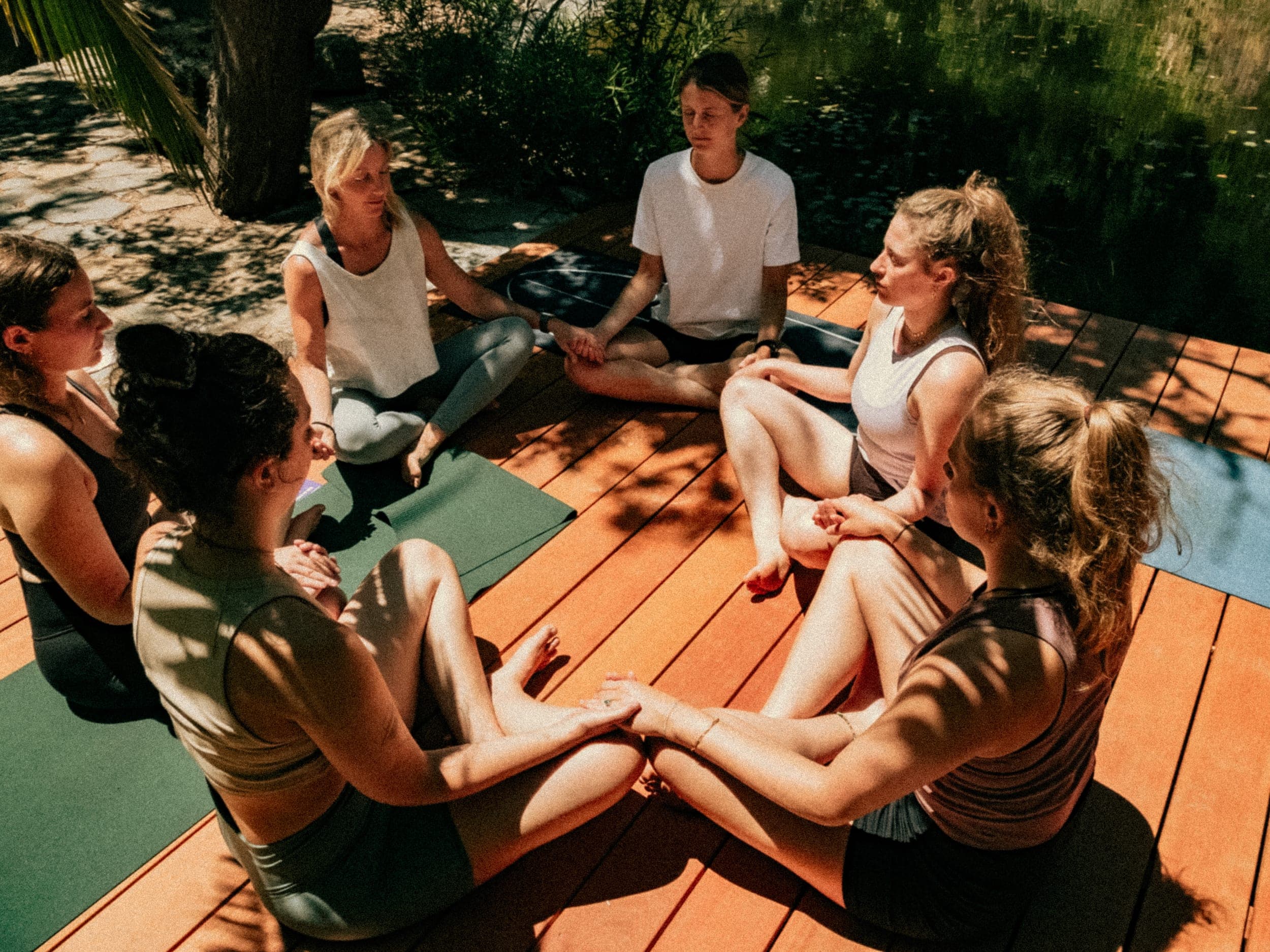 Group of women sitting cross-legged in a circle meditating on a sunlit wooden deck