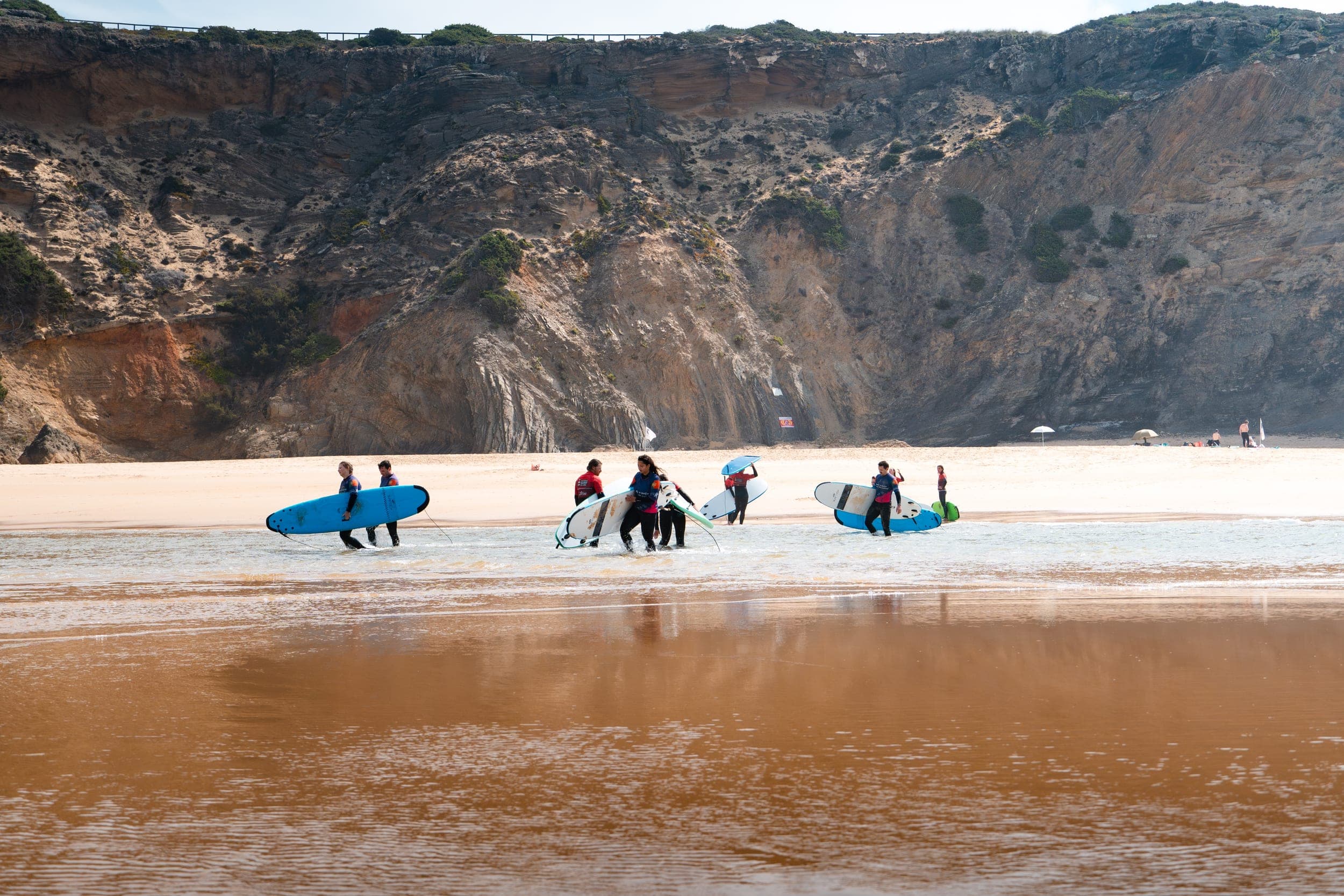 Surf group carrying boards across a shallow river below towering Algarve cliffs
