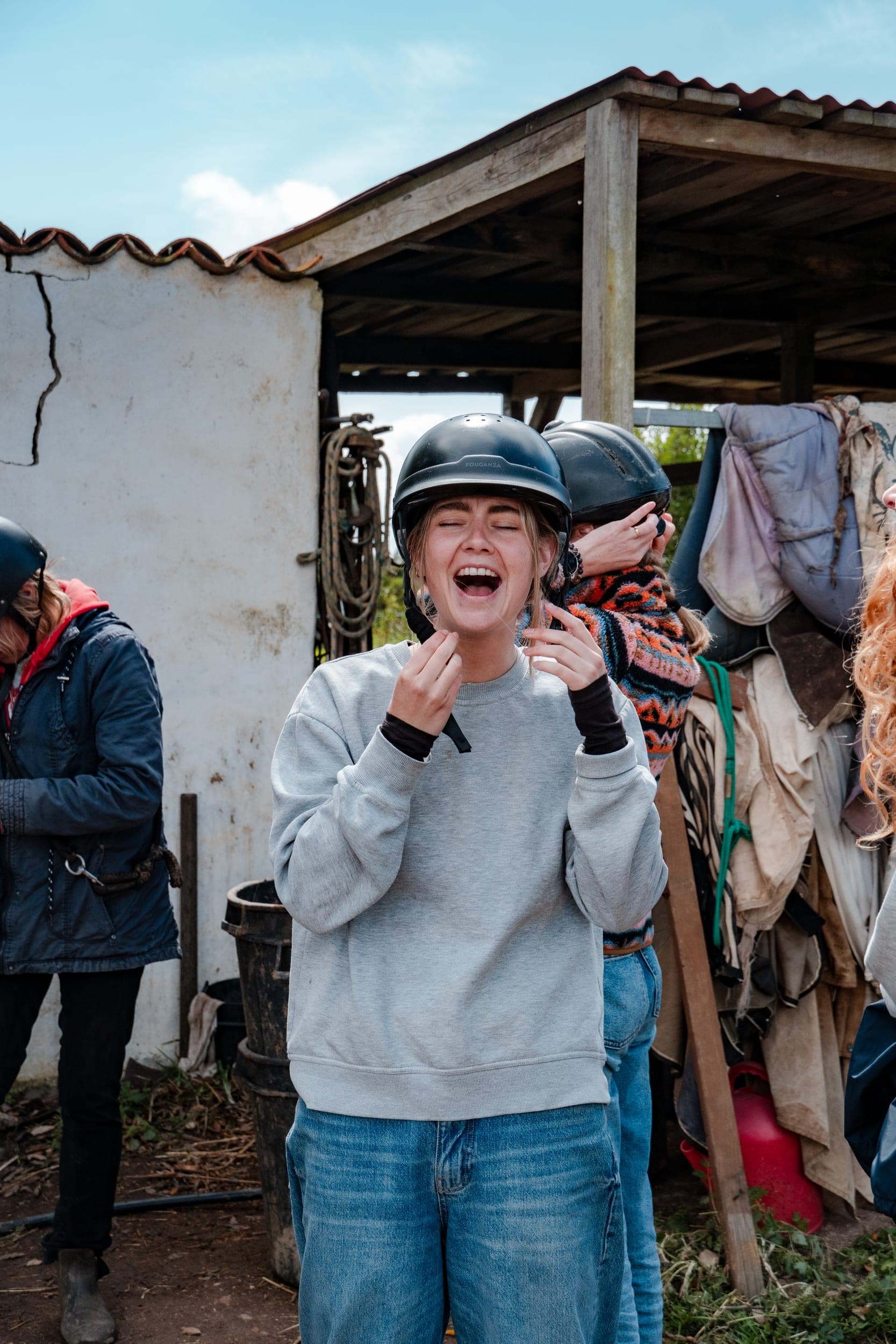 Laughing woman fastening her riding helmet outside a rustic stable with tack