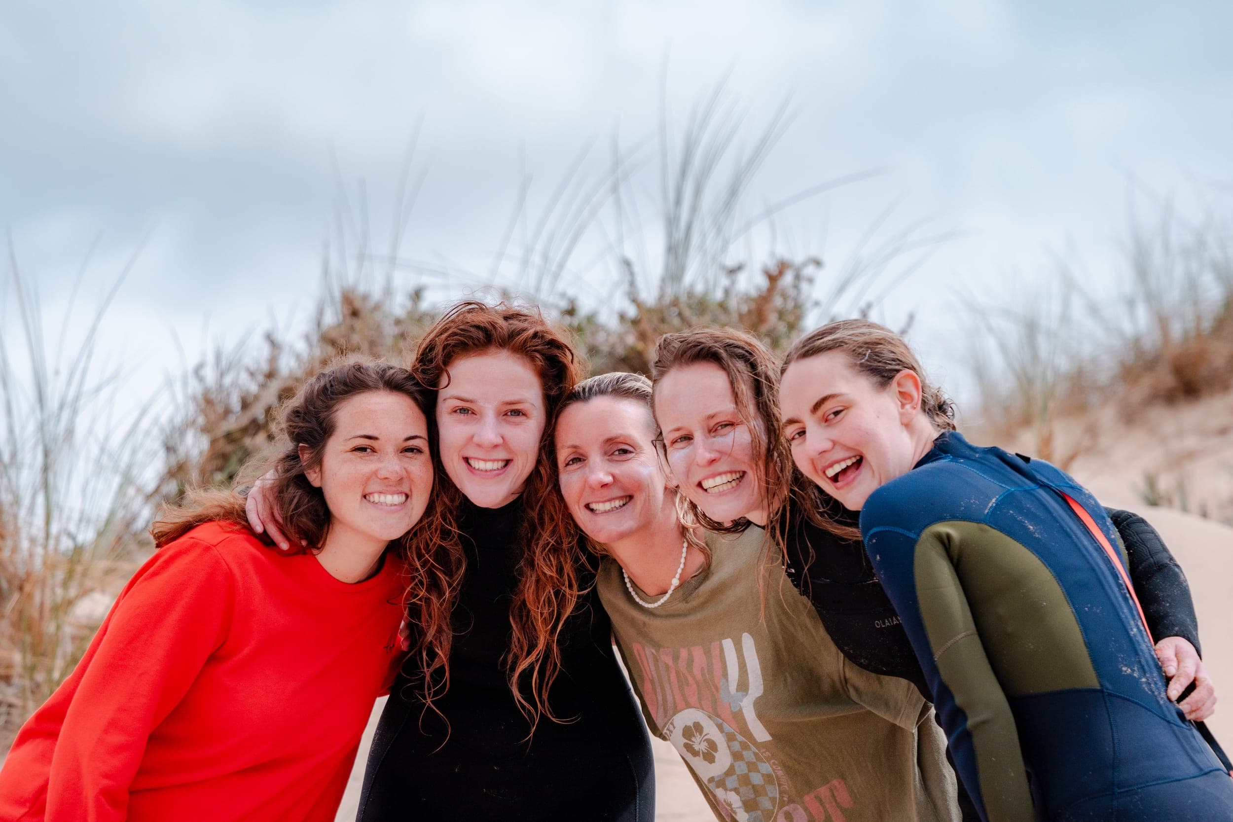 Five women in wetsuits smiling cheek to cheek on a windswept beach dune