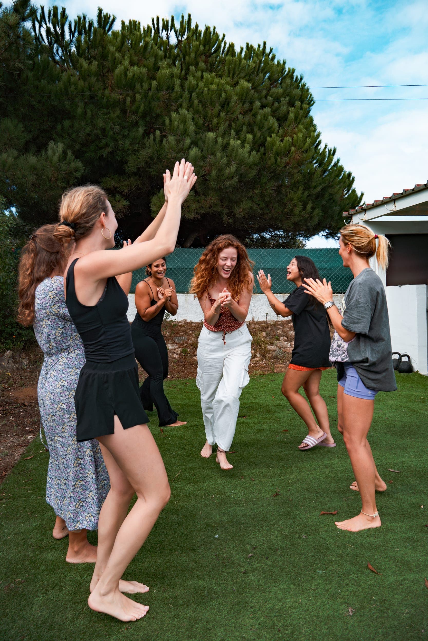 Group of women laughing and clapping while dancing barefoot on grass under a pine tree