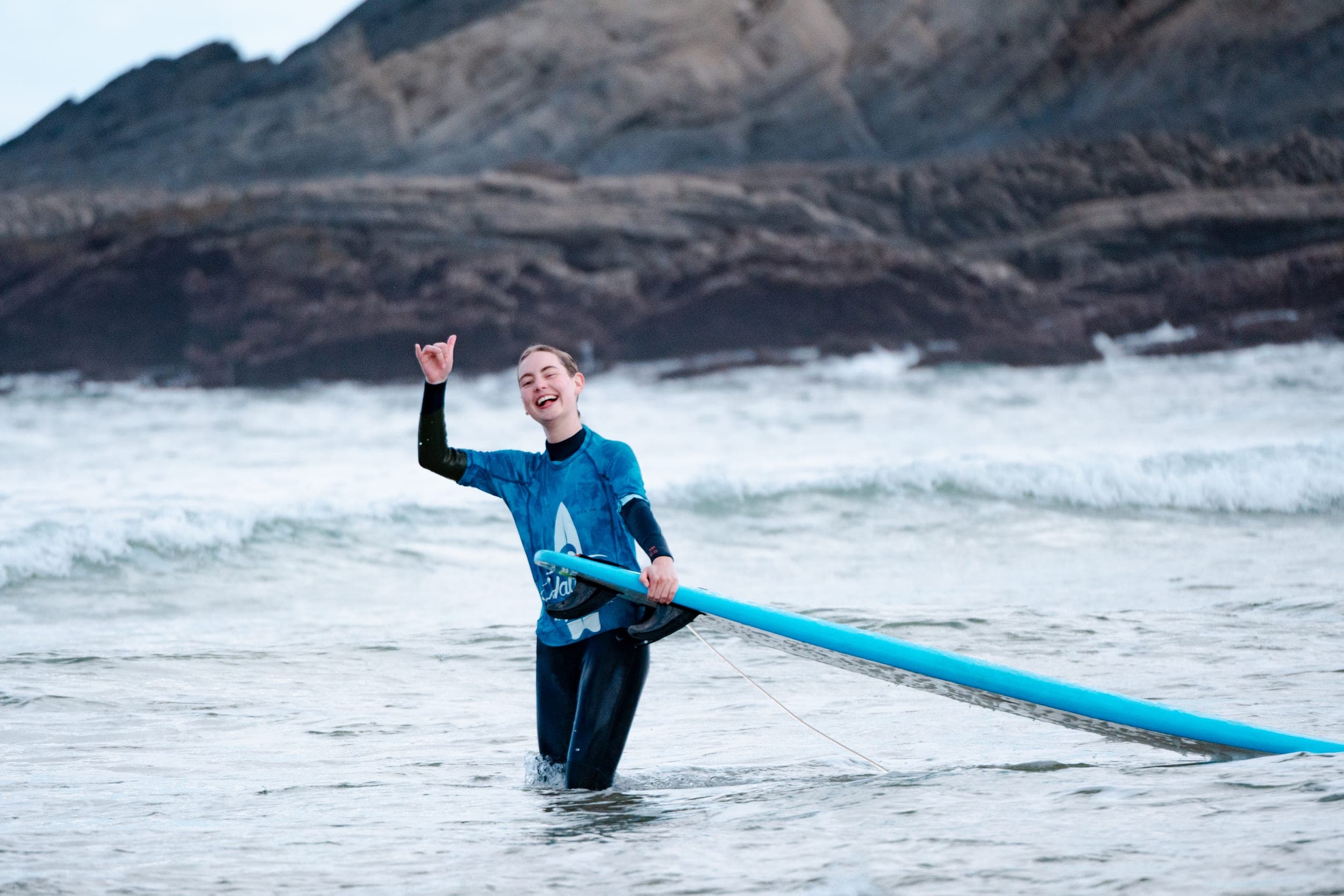 Woman in blue rash vest laughing and throwing a shaka while standing in the surf