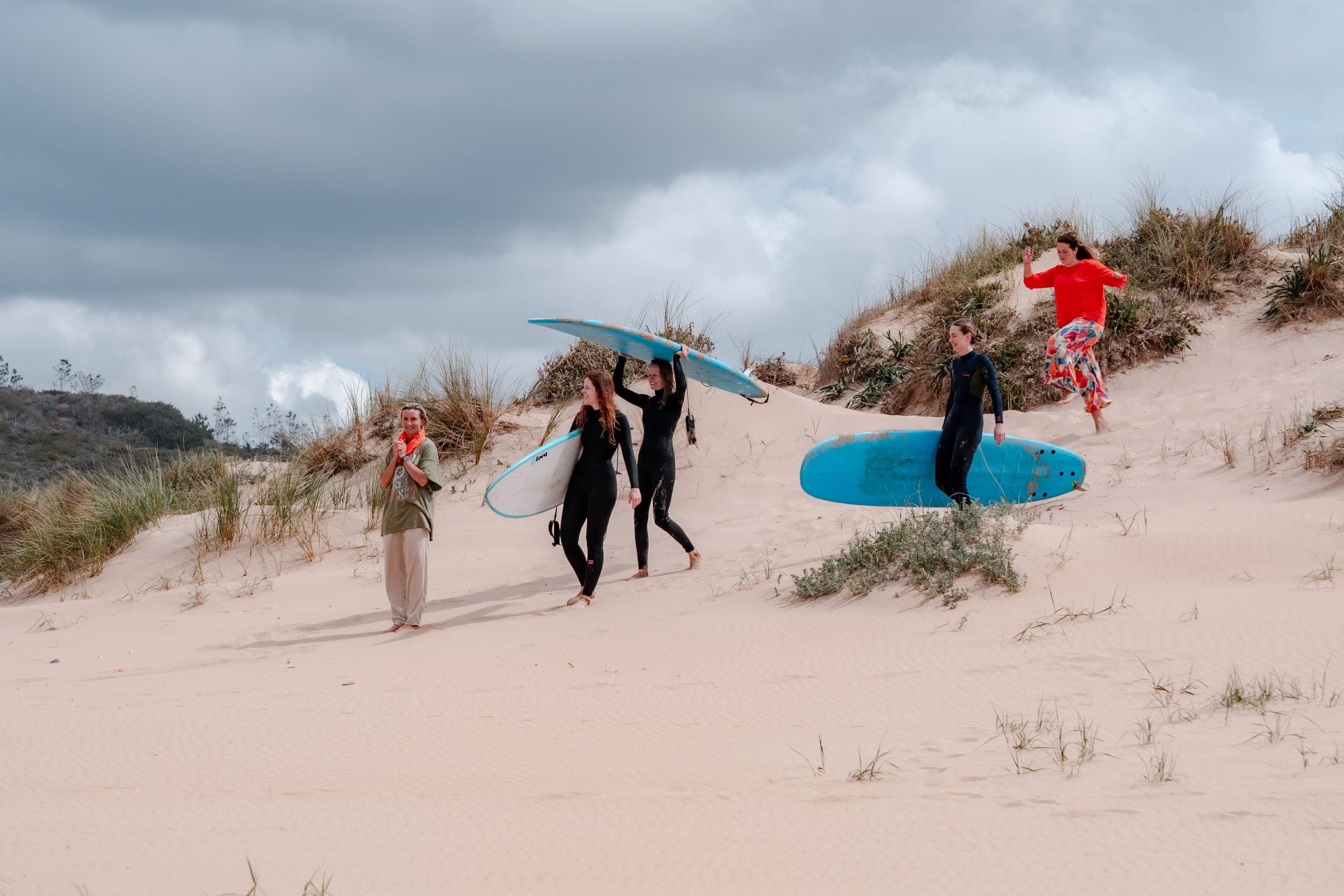 Women in wetsuits carrying surfboards down a sandy dune under a stormy grey sky