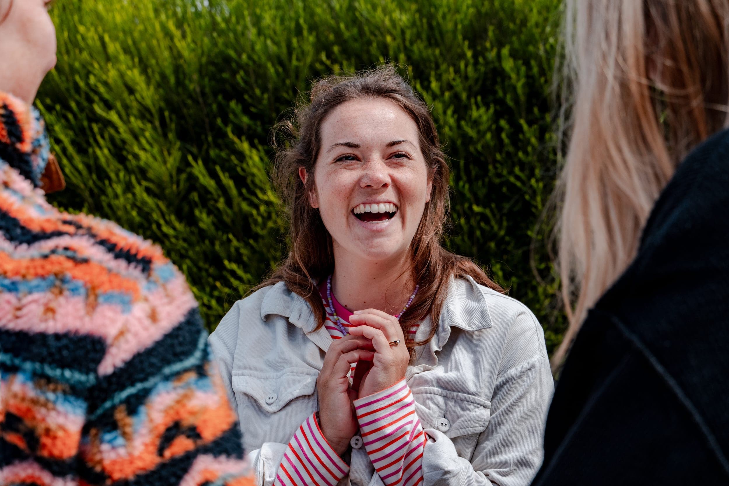 Woman in a corduroy jacket laughing warmly during a conversation by a green hedge