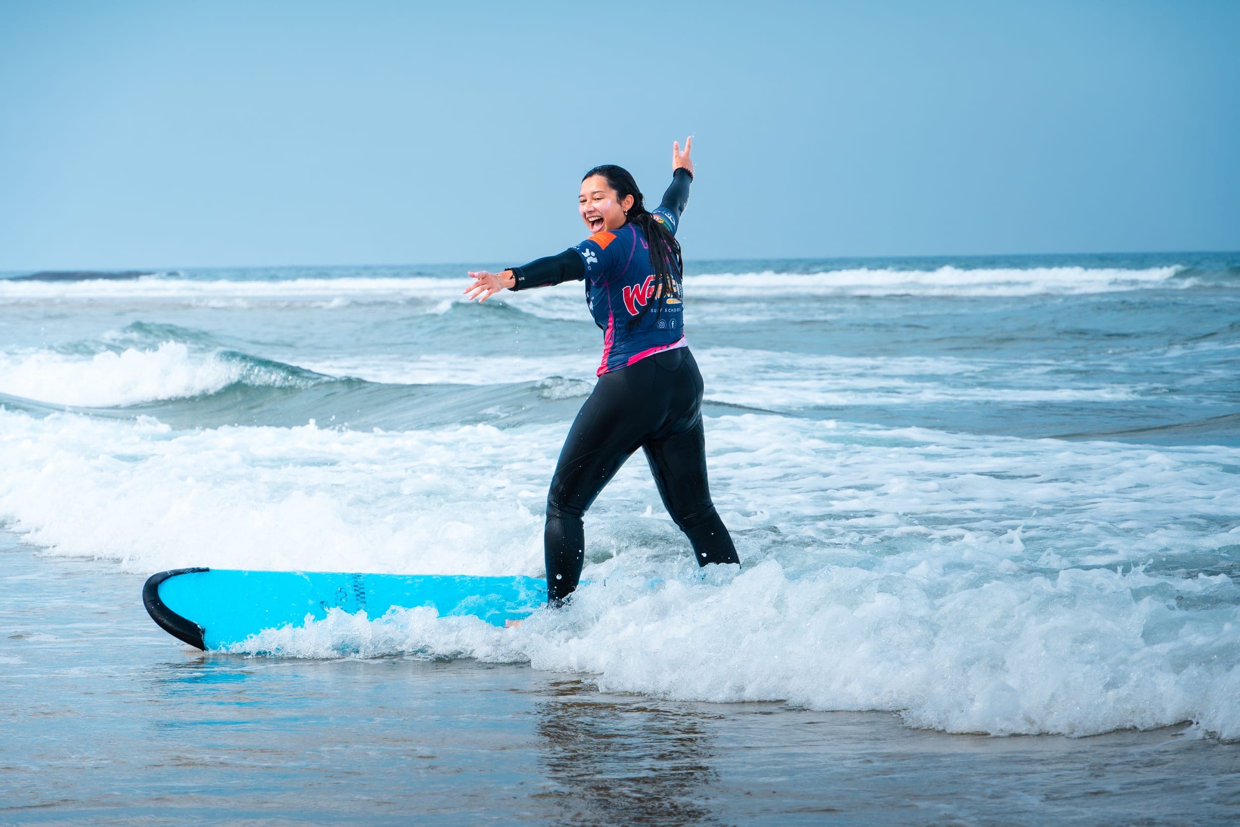 Woman in wetsuit riding a blue surfboard through whitewater with arms spread wide