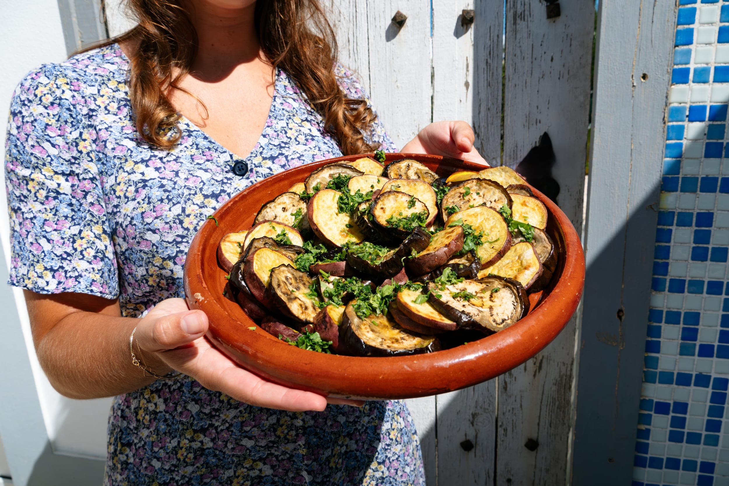 Woman in a floral dress holding a clay dish of roasted eggplant and sweet potato with herbs