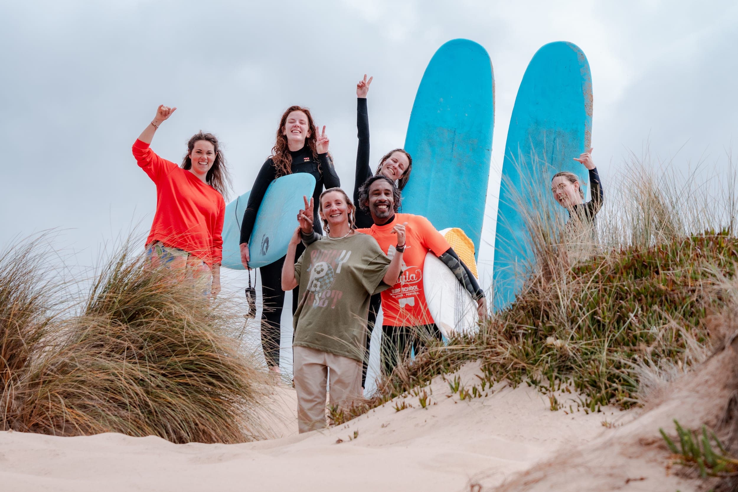 Six friends posing with surfboards on a grassy dune, throwing shakas and peace signs