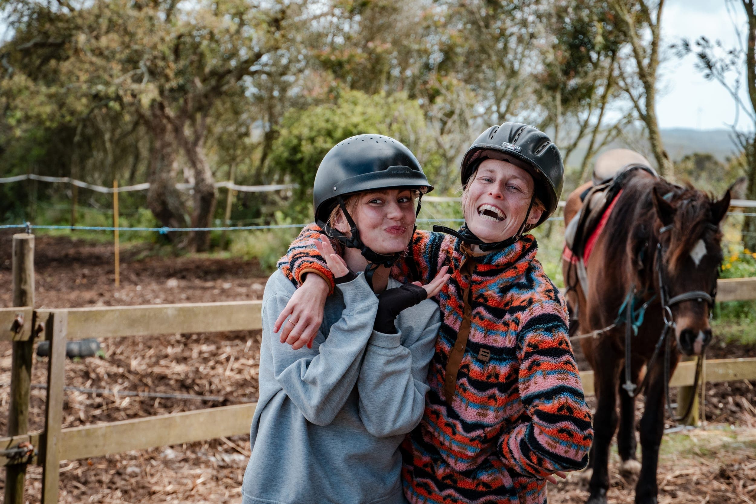 Two women in riding helmets laughing and hugging beside a brown horse at a paddock