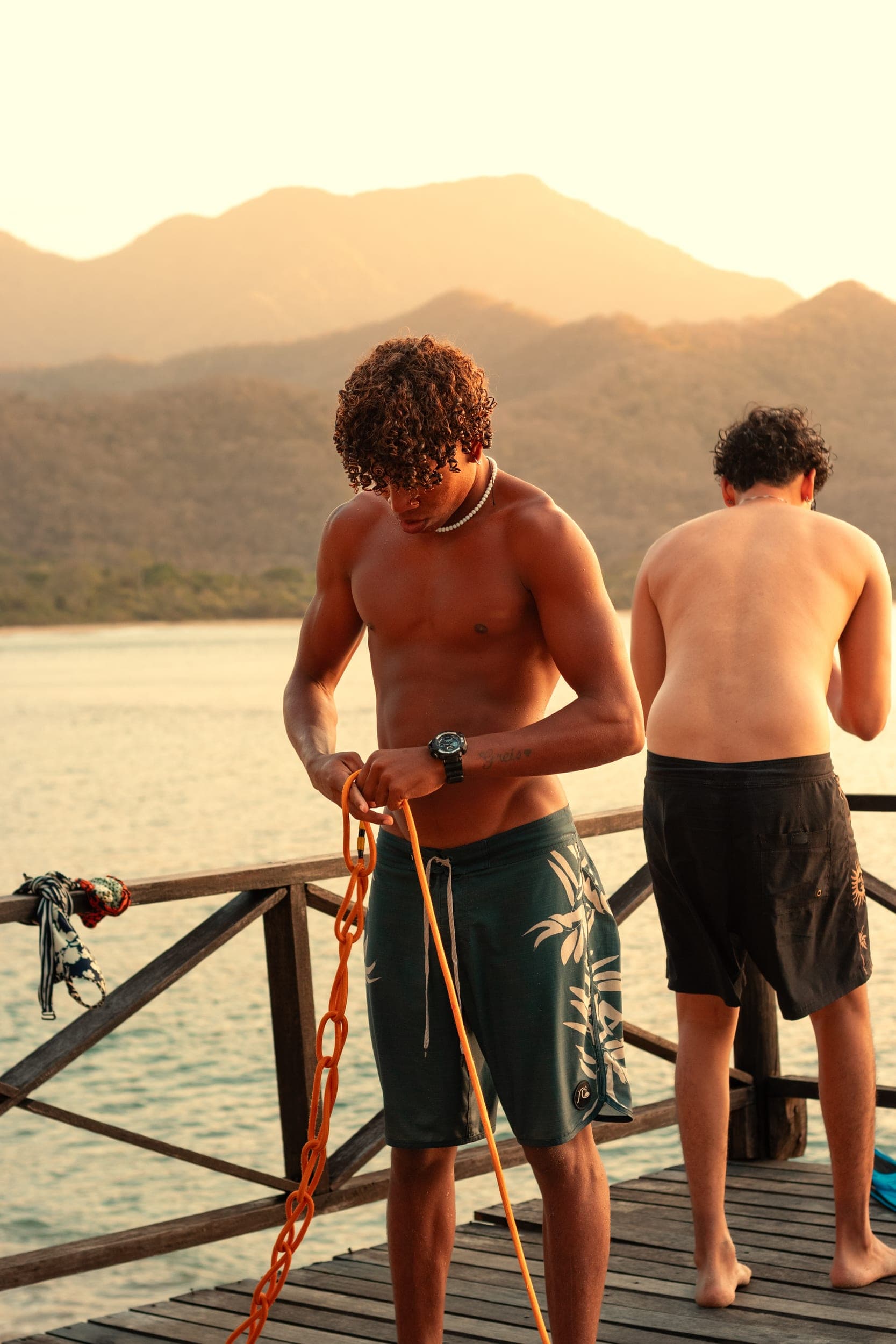 Shirtless man coiling rope on a jetty at golden hour with mountains across the bay