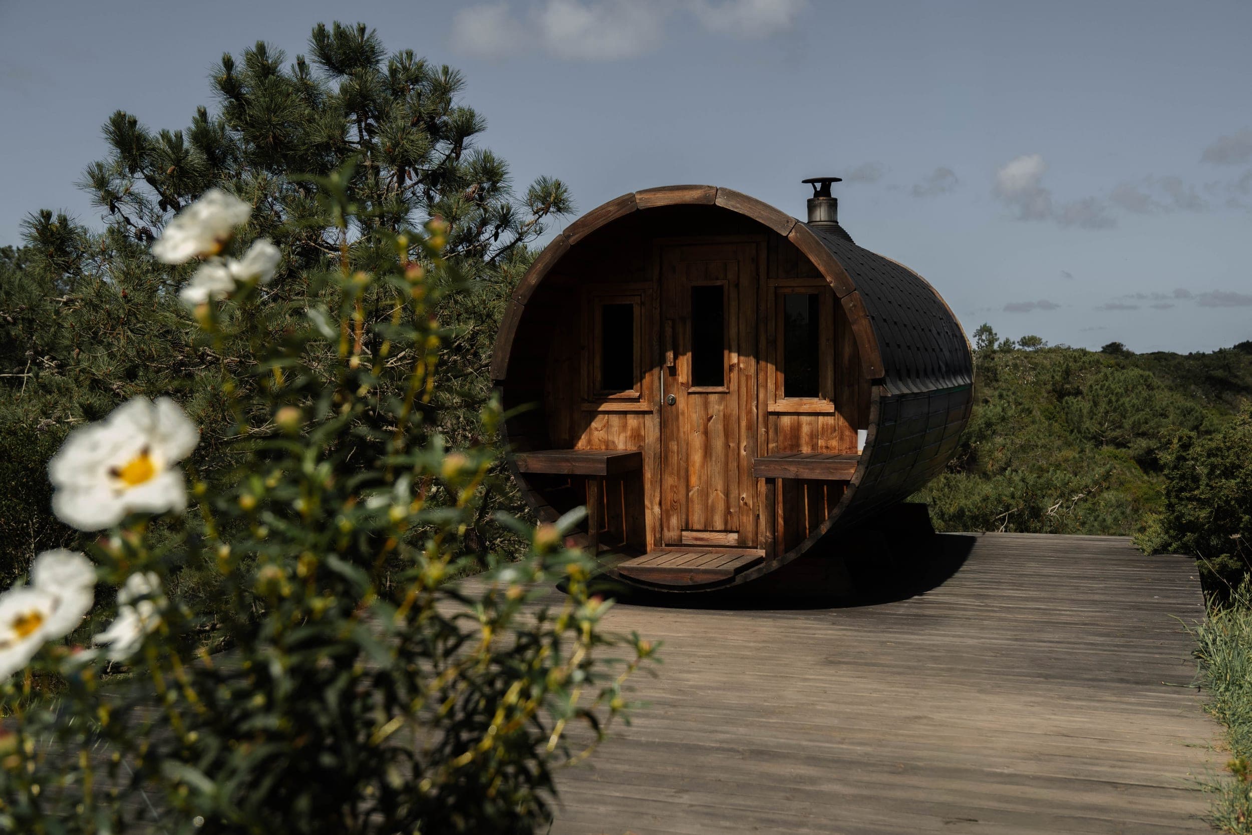 Wooden barrel sauna on a deck framed by pine and white rockrose flowers