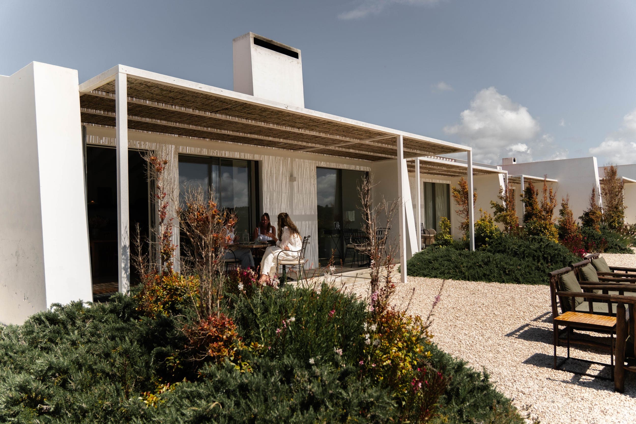 Two women seated at a terrace table under a villa pergola surrounded by garden shrubs