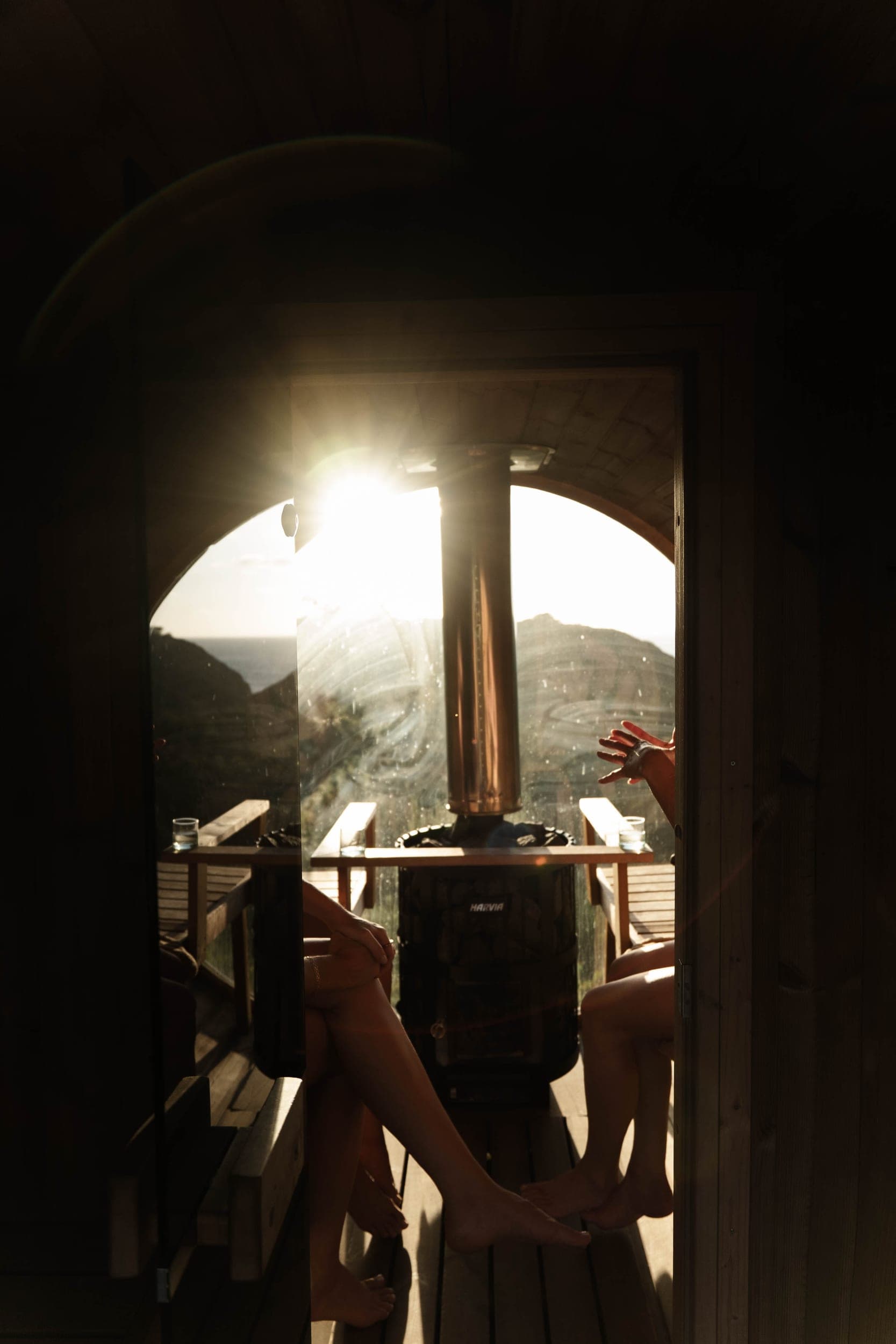 Two women's legs inside a barrel sauna backlit by a blazing sunset over the coast