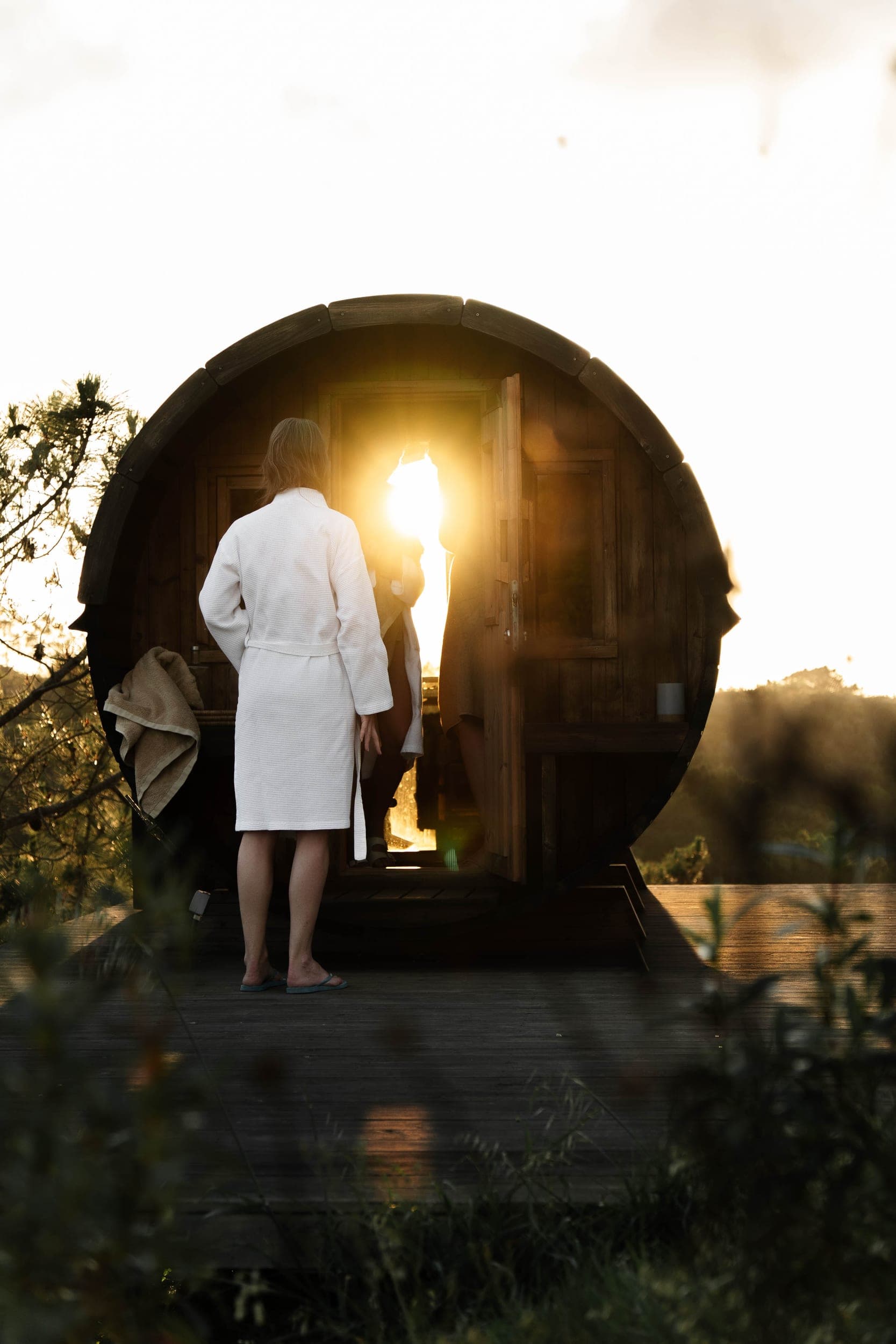 Woman in a white robe entering a barrel sauna glowing with sunset light on a deck