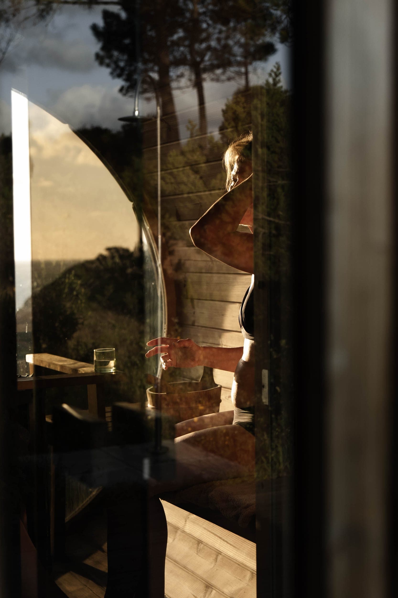 Woman in a swimsuit inside a barrel sauna seen through reflective glass at golden hour