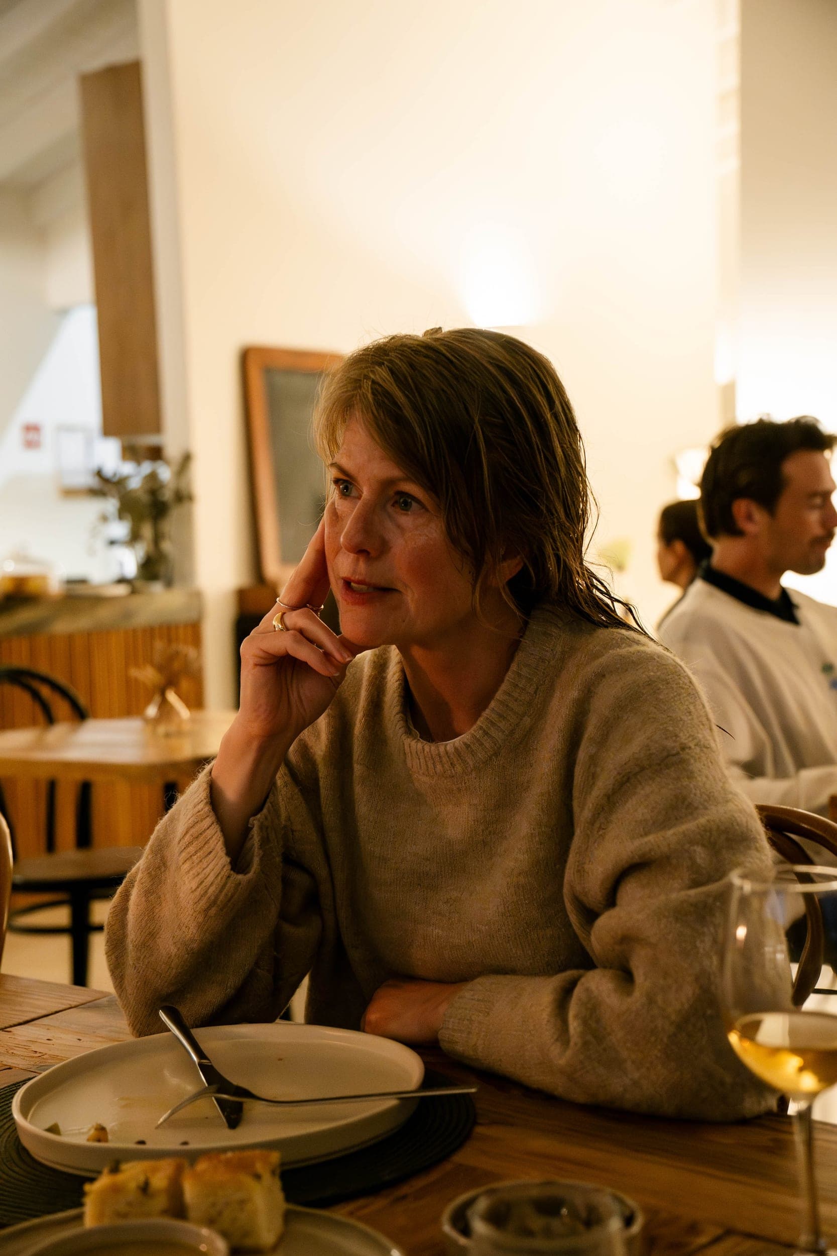 Woman in a beige sweater resting her chin on her hand at a restaurant table, wine nearby