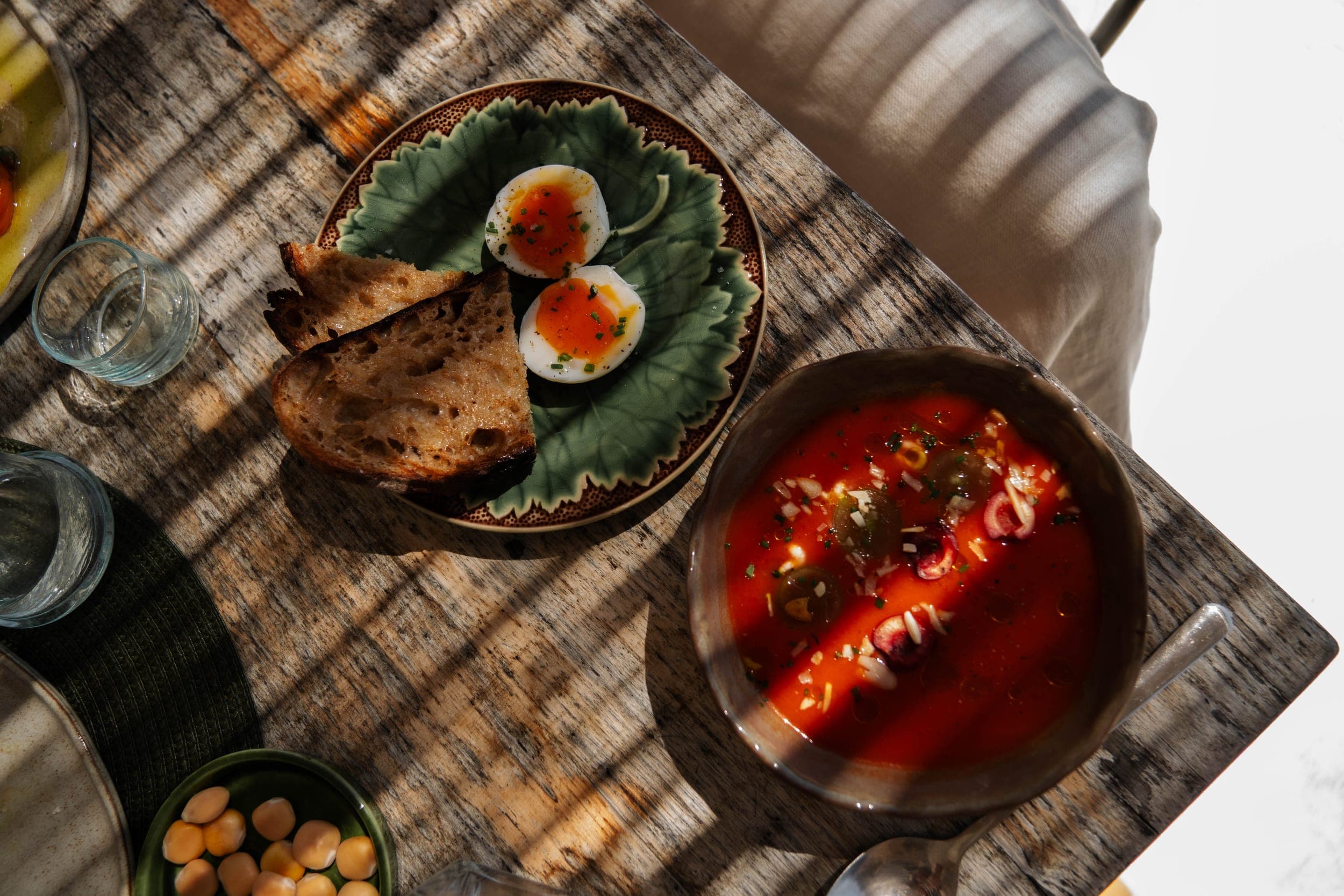 Overhead brunch of soft-boiled eggs on a leaf plate, sourdough toast and tomato soup in dappled sun
