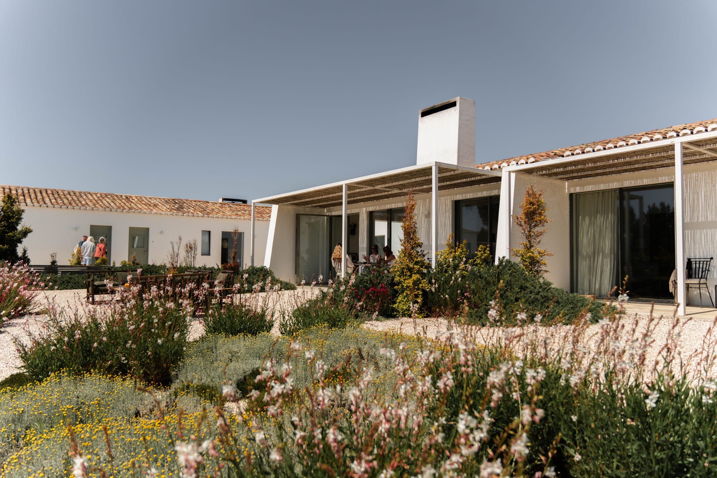 Guests relaxing on a white villa's pergola terrace above a flowering meadow garden
