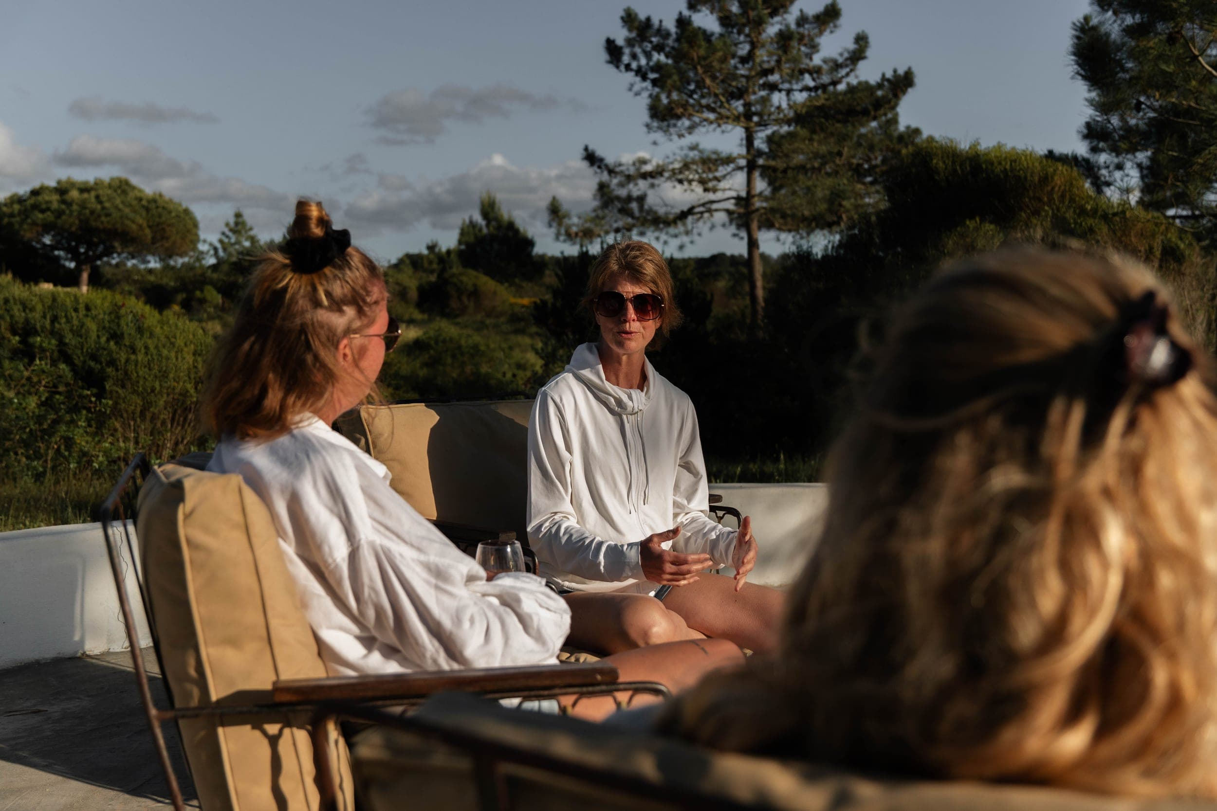 Three women in white chatting on a sunlit patio at golden hour with woodland behind