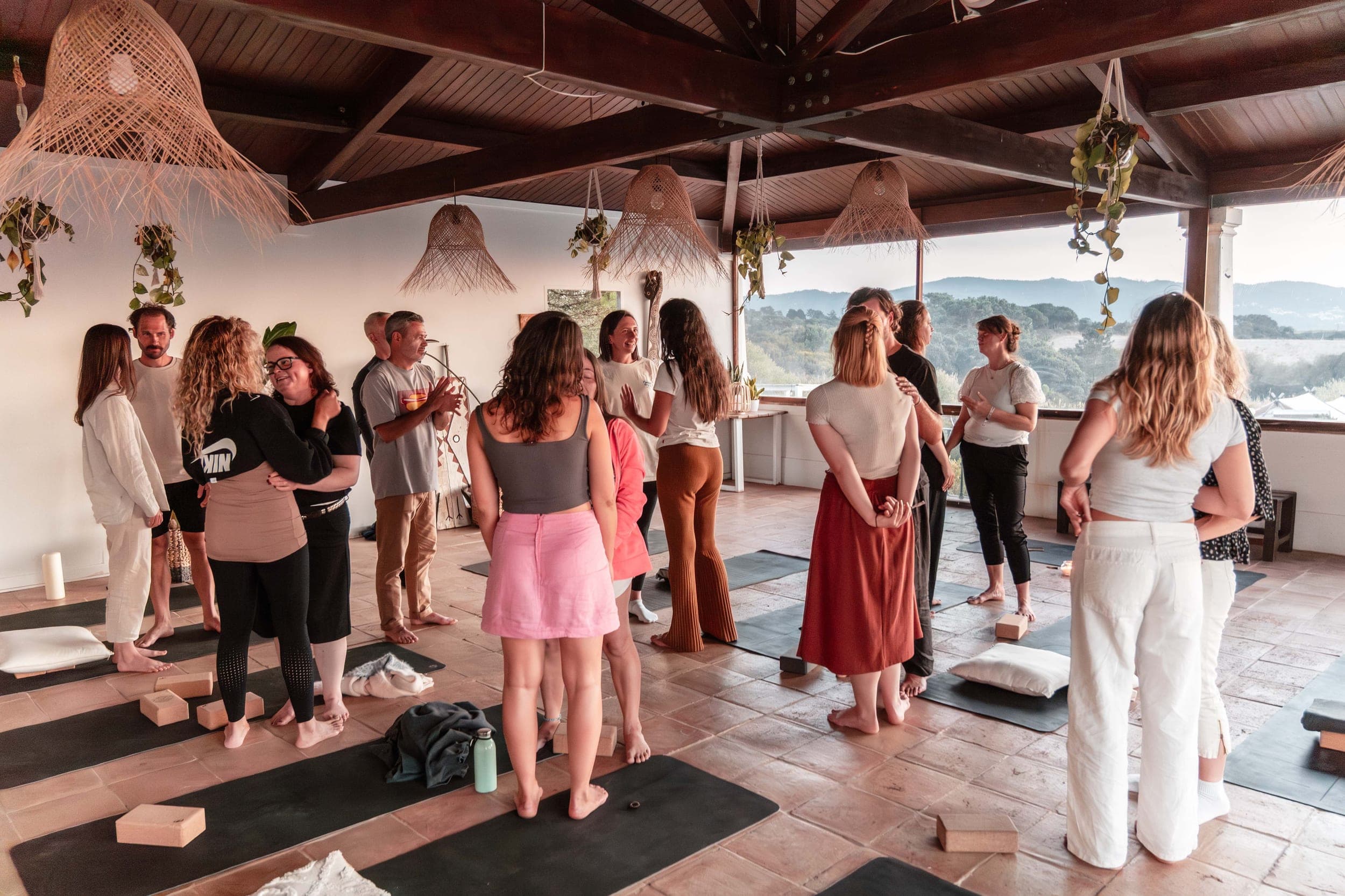 Group standing in a circle chatting on yoga mats in a sunlit studio with hill views