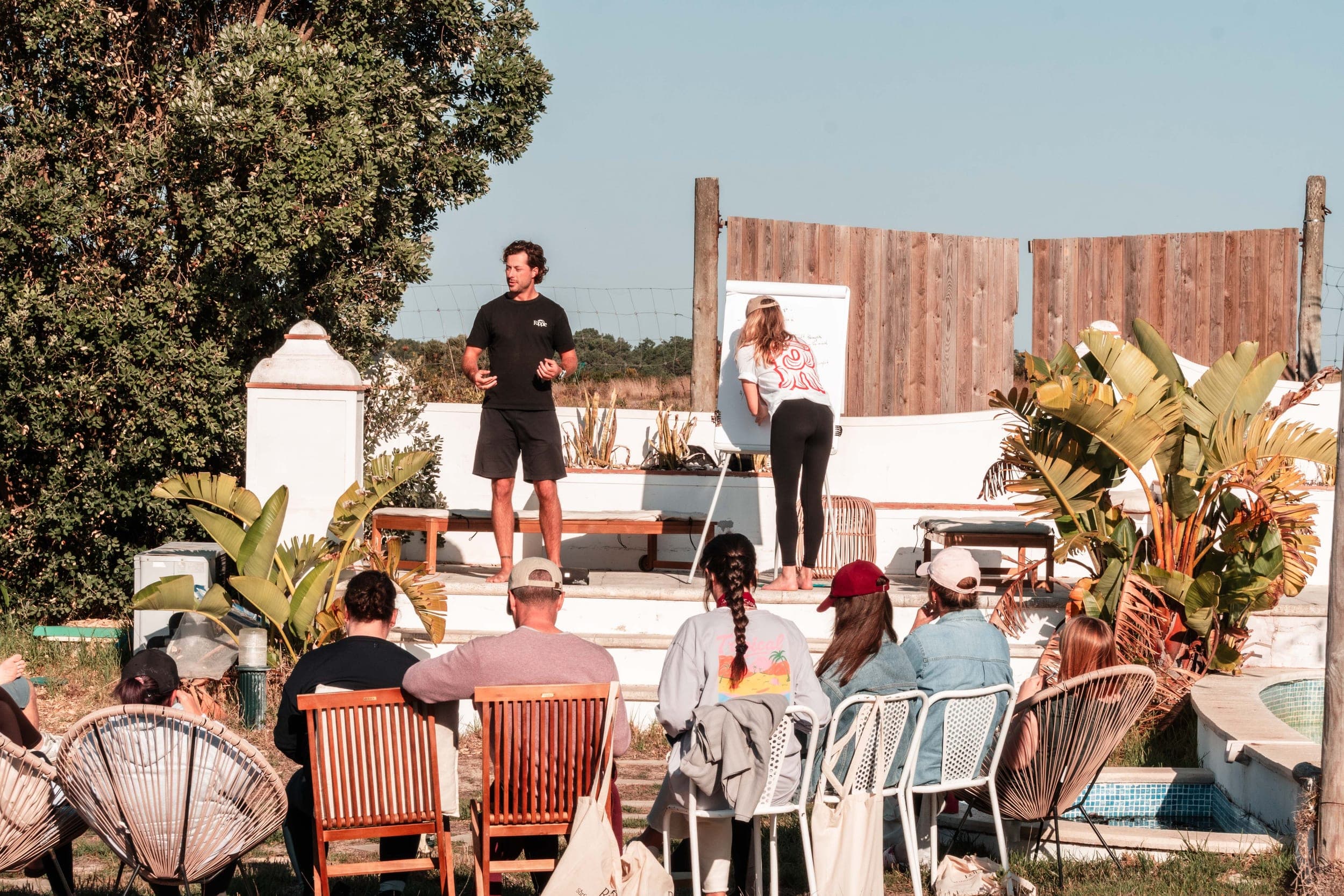 Instructor speaking to a seated group beside a flip chart at an outdoor poolside workshop