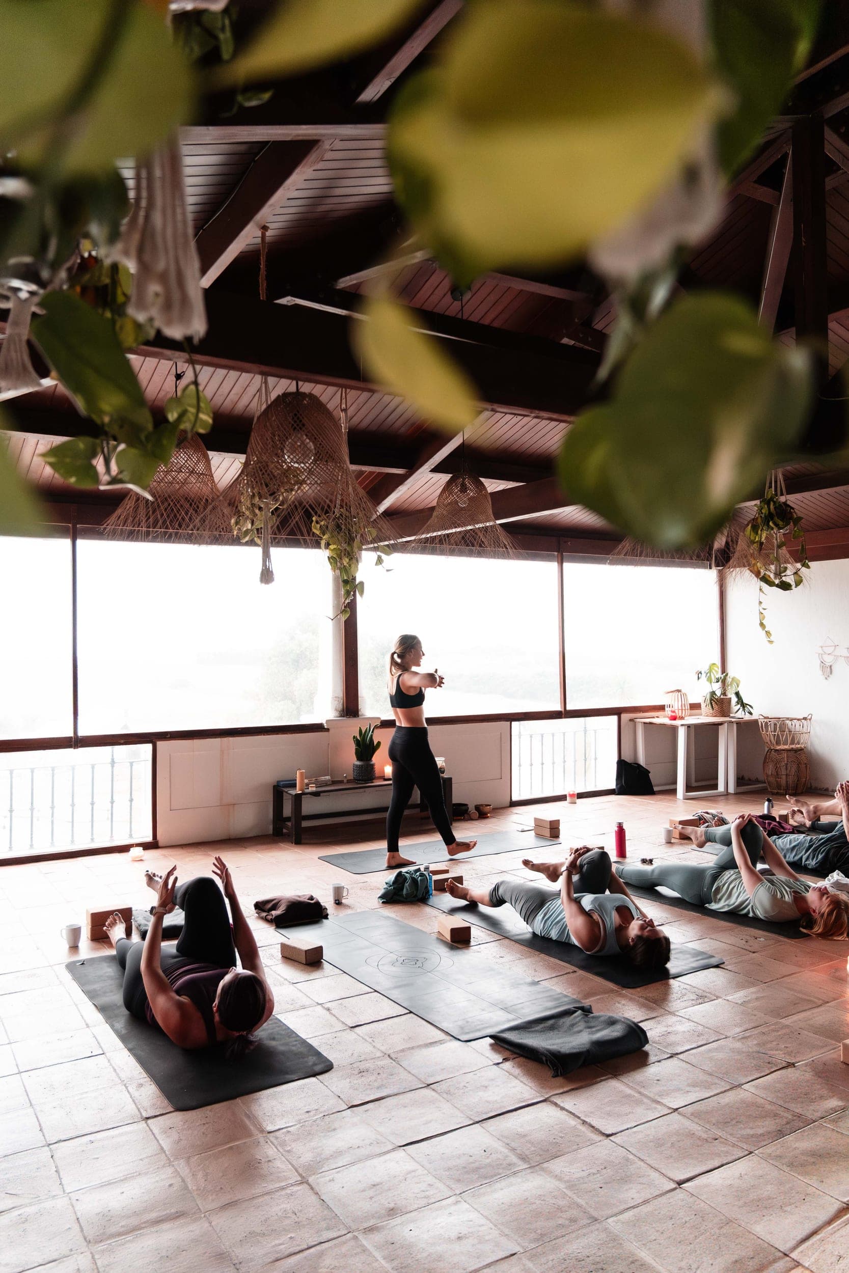 Woman leading a stretching yoga class as participants lie on mats in a plant-filled studio