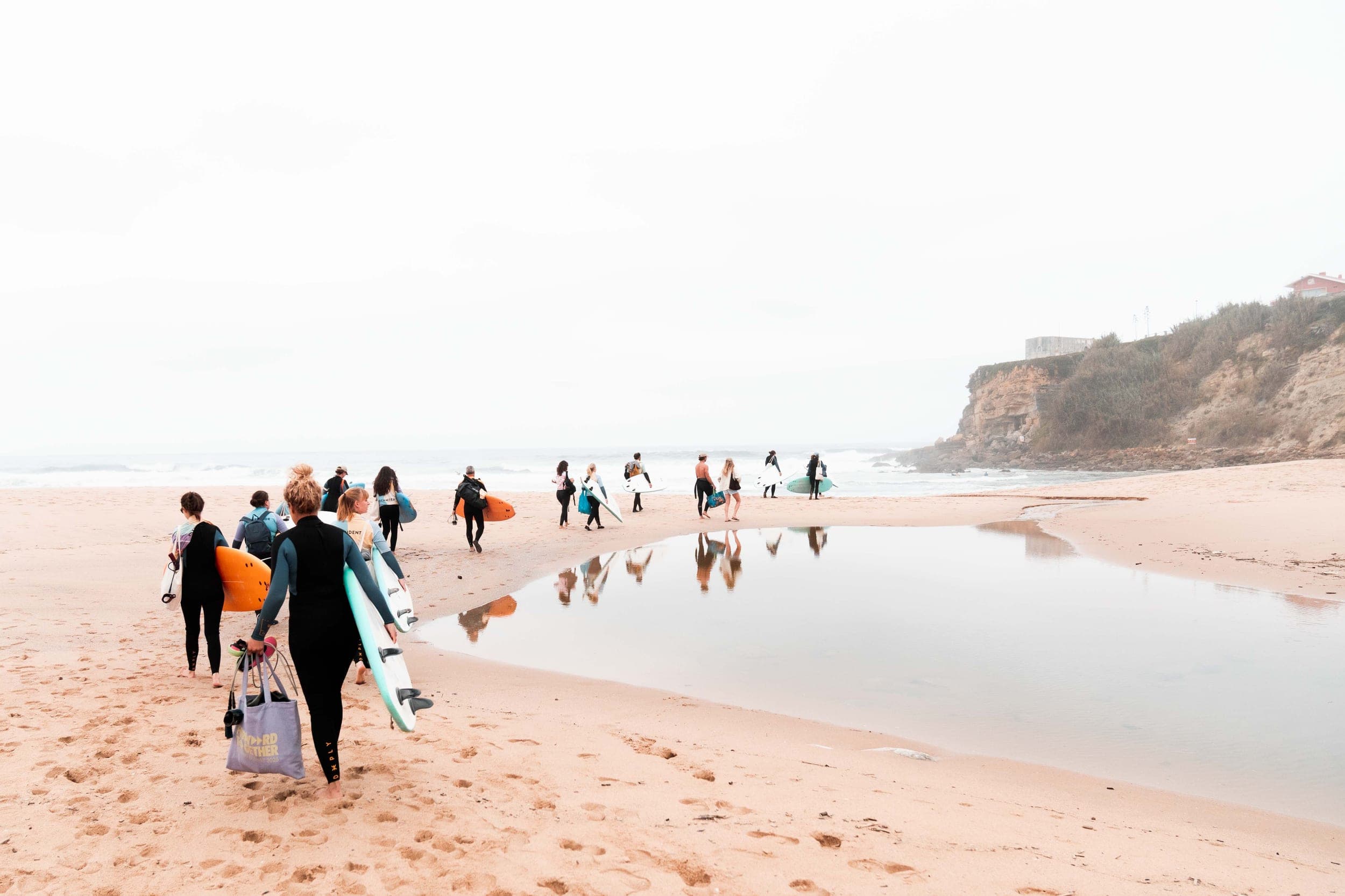 Group in wetsuits carrying surfboards across the beach toward the sea and cliffs