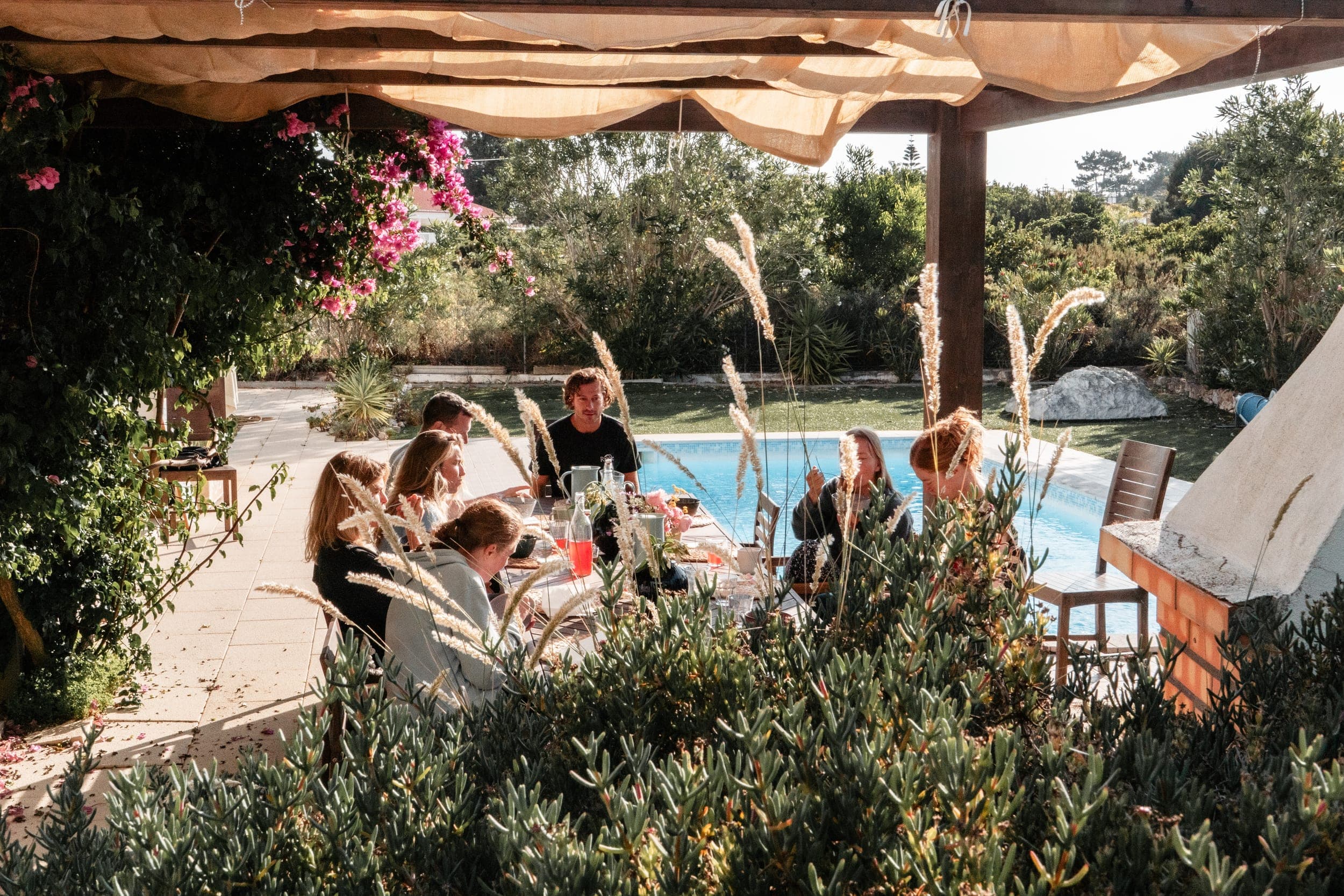 Guests sharing a meal at a long poolside table framed by garden flowers and greenery