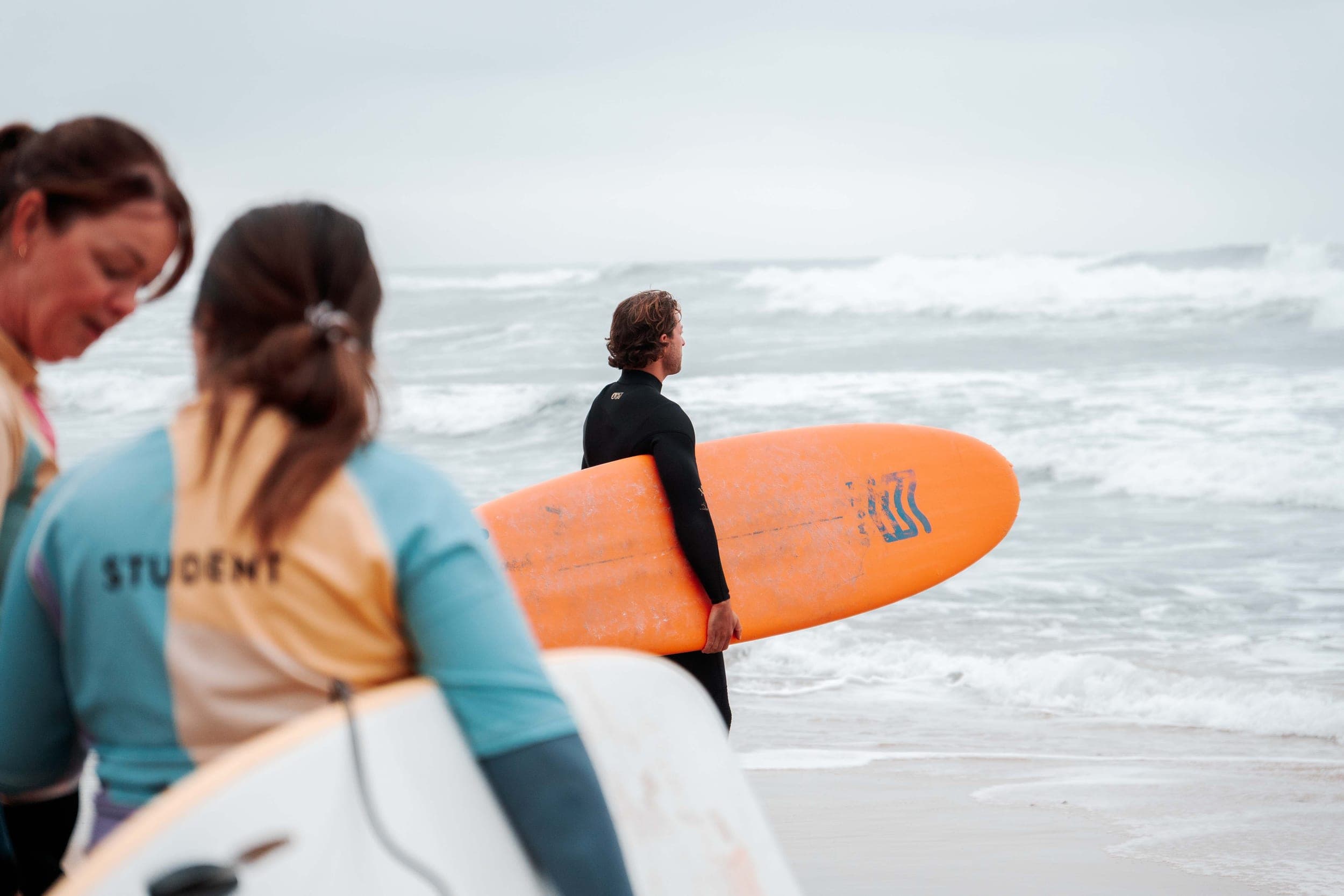 Surfer in wetsuit carrying an orange board toward the waves as two students look on
