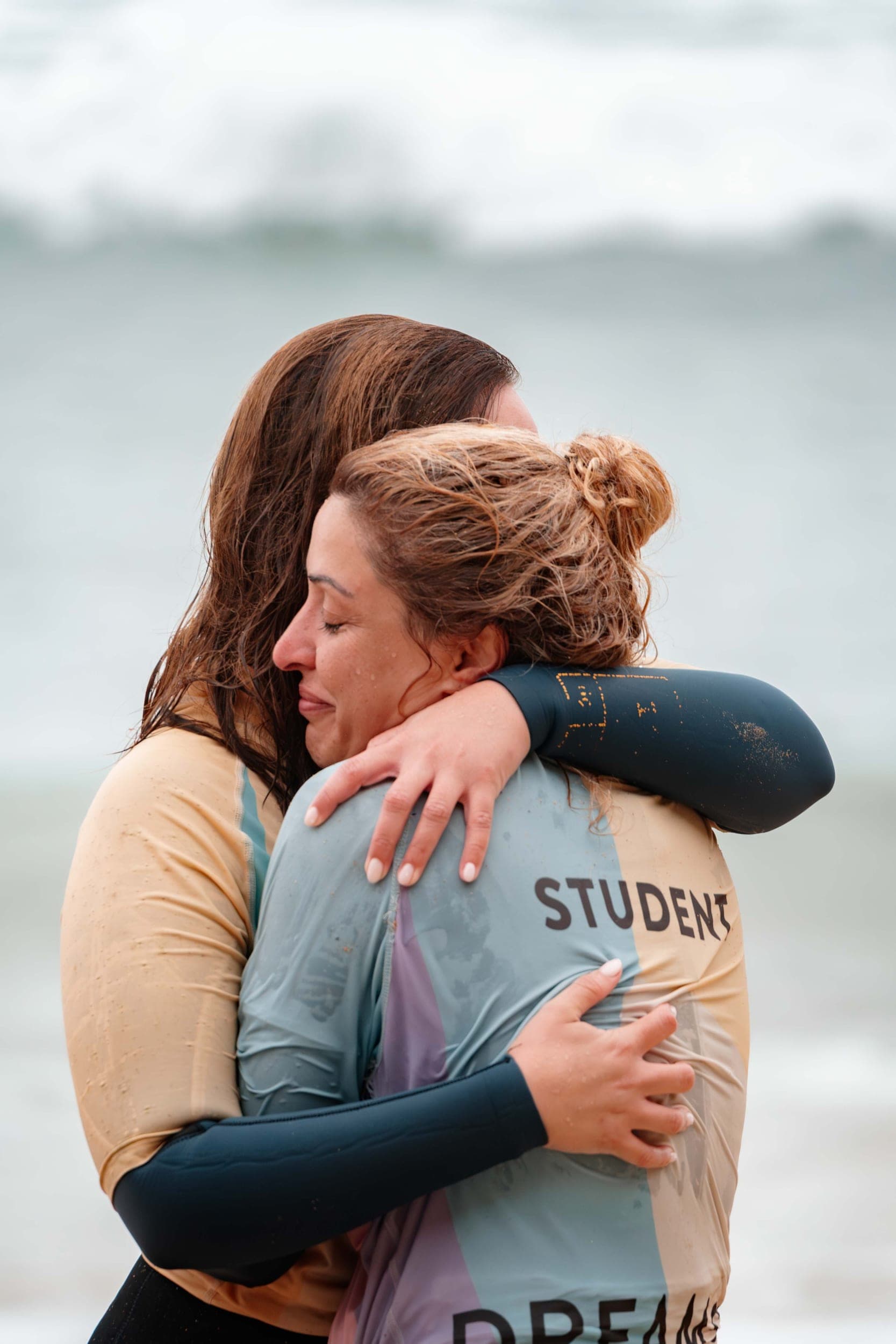 Two women in wetsuits sharing an emotional embrace on the beach, one wearing a student rash vest