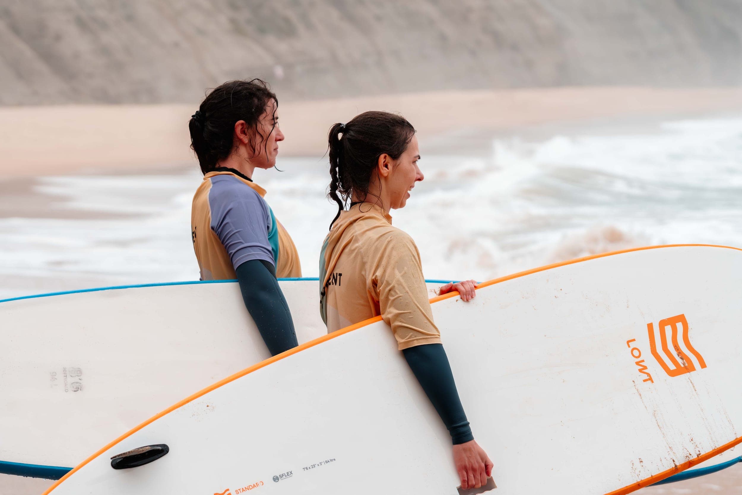 Two women in rash vests holding surfboards, gazing at the sea below sandy cliffs