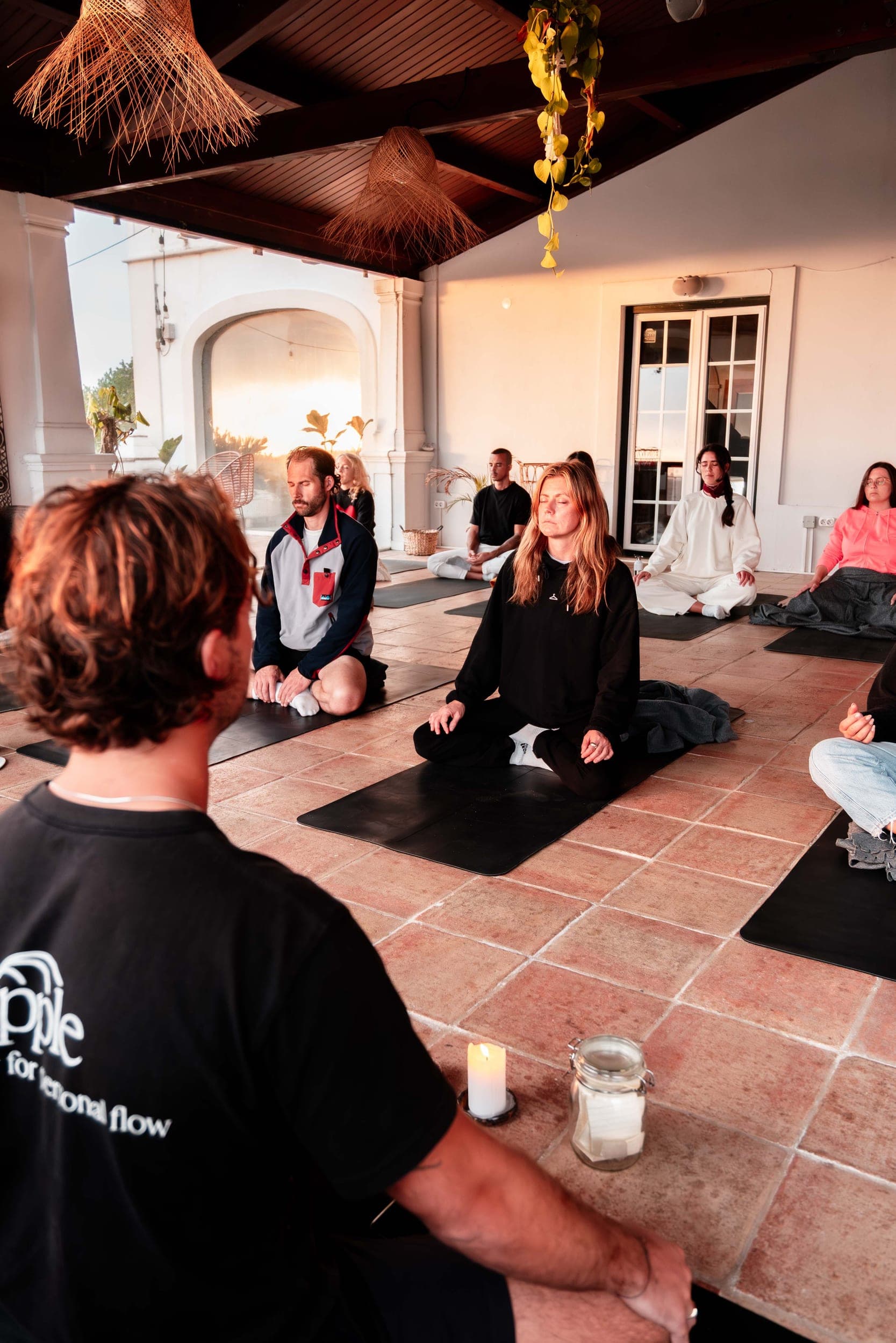Group meditating cross-legged on mats during a candlelit session on a sunset patio