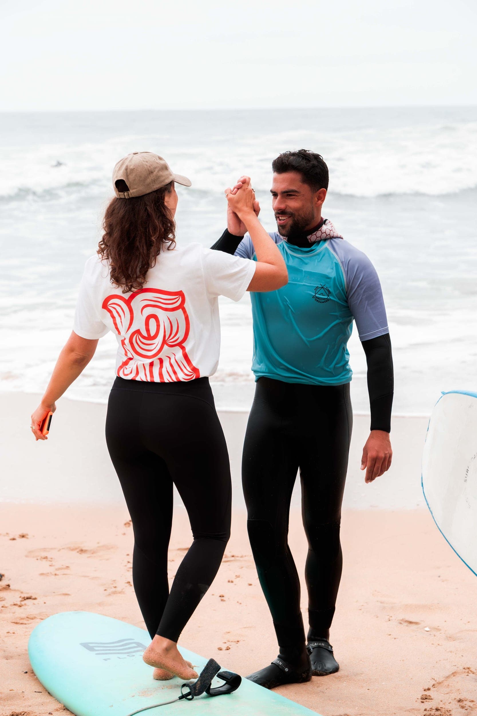 Woman and male instructor in wetsuits high-fiving on the beach beside a surfboard