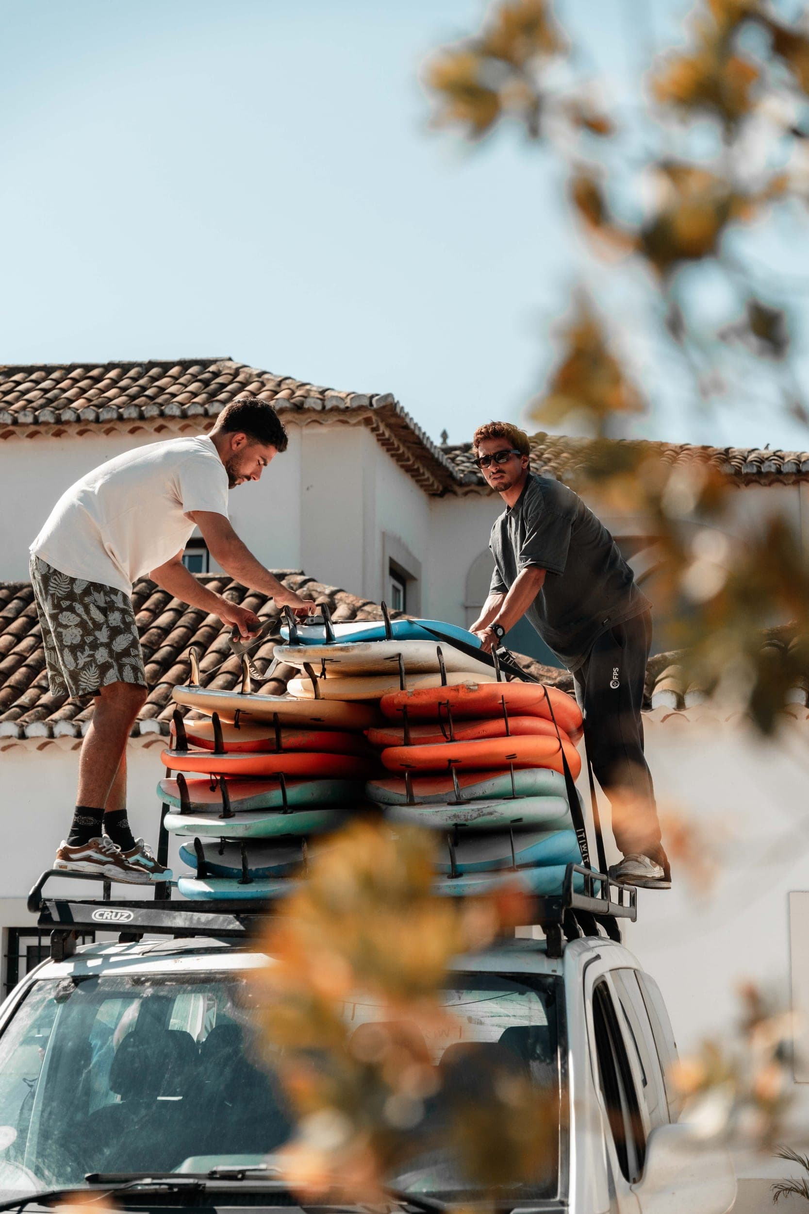 Two men strapping a stack of surfboards onto a car roof rack outside a tiled-roof villa