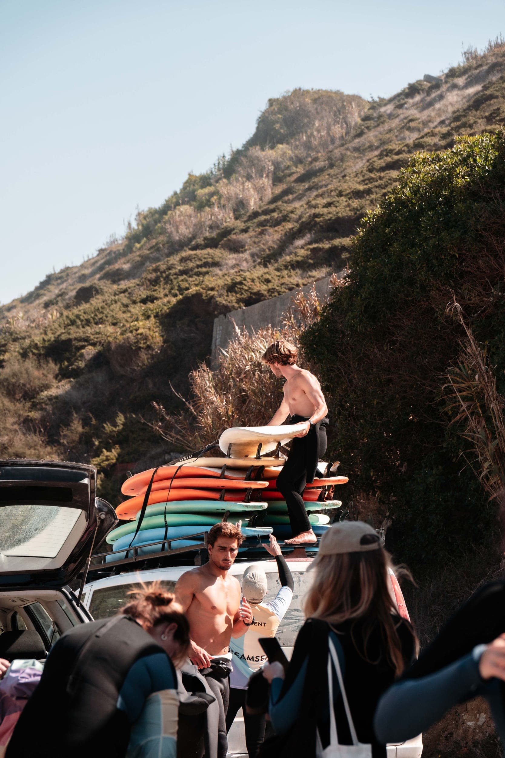 Shirtless surfer unloading colorful surfboards from a car roof rack below a green hillside