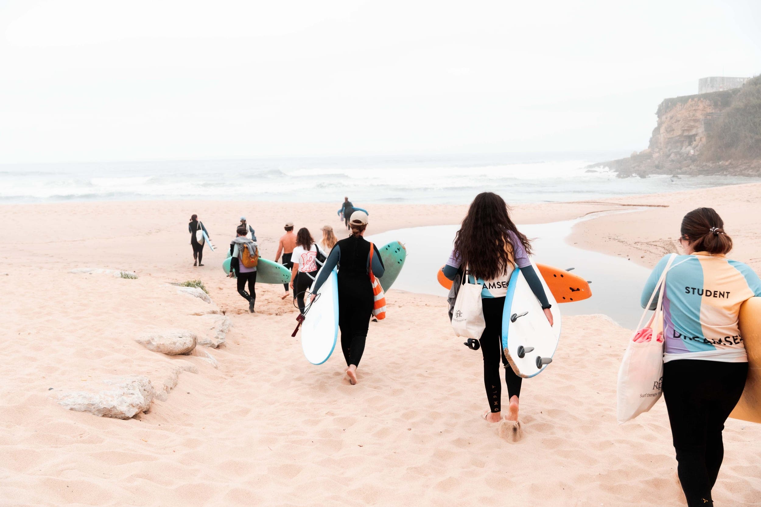 Wetsuit-clad group carrying surfboards down a sandy beach toward the ocean and cliffs
